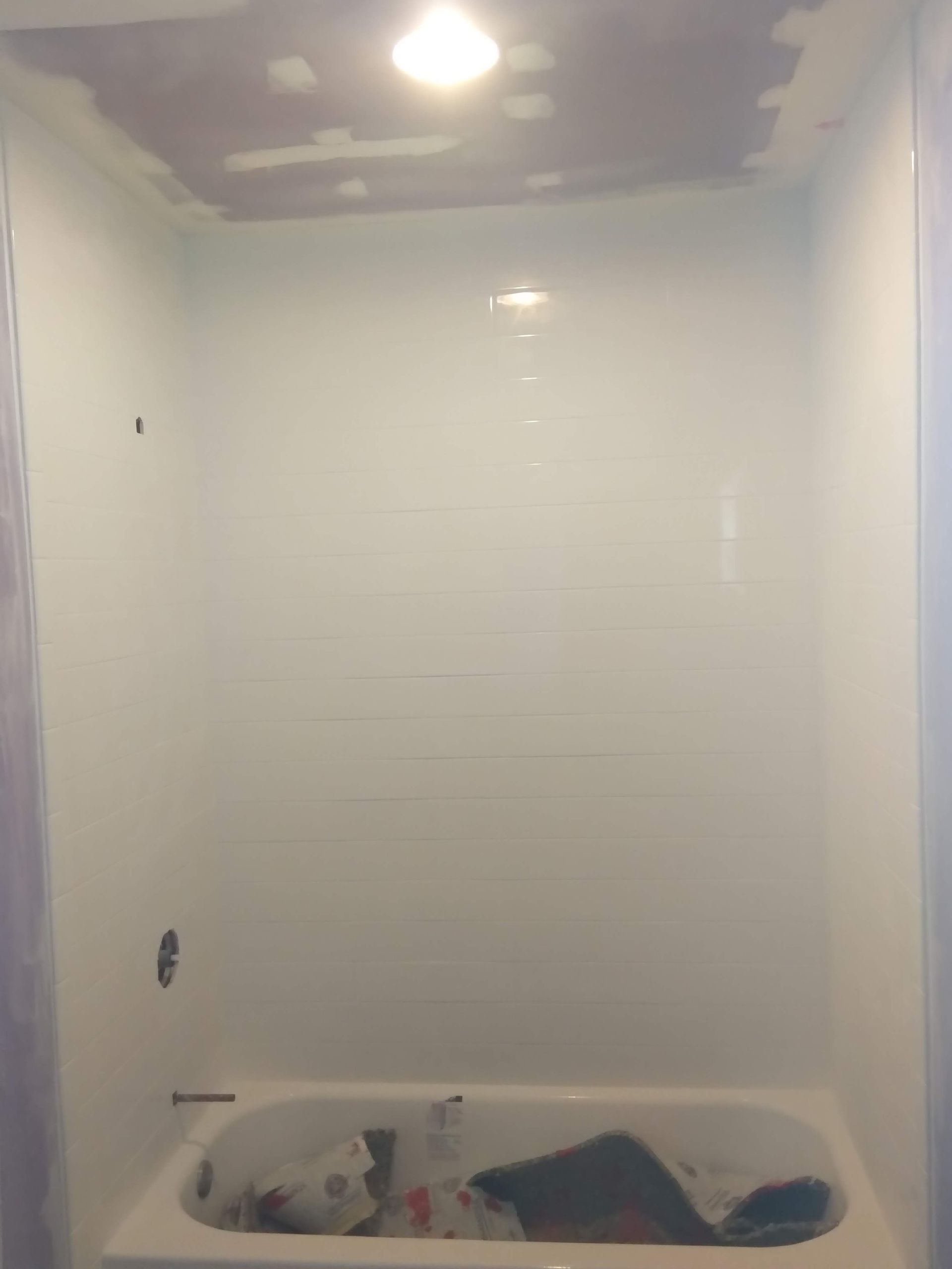 White shower/tub stall with a bare wall, ceiling light, and a cluttered tub floor with paint cans and debris