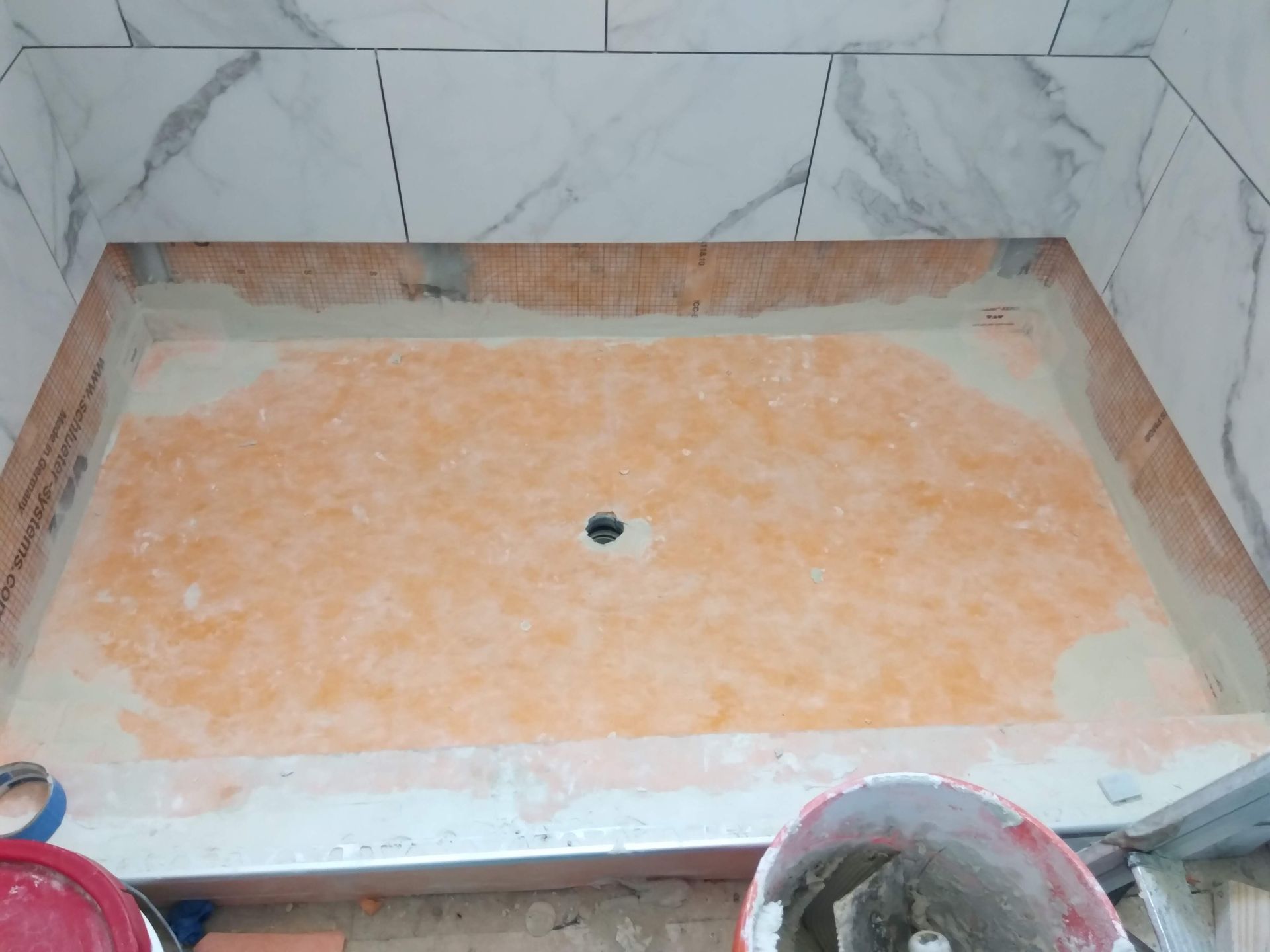 Empty tiled shower floor with a central drain and a pale orange waterproofing layer.