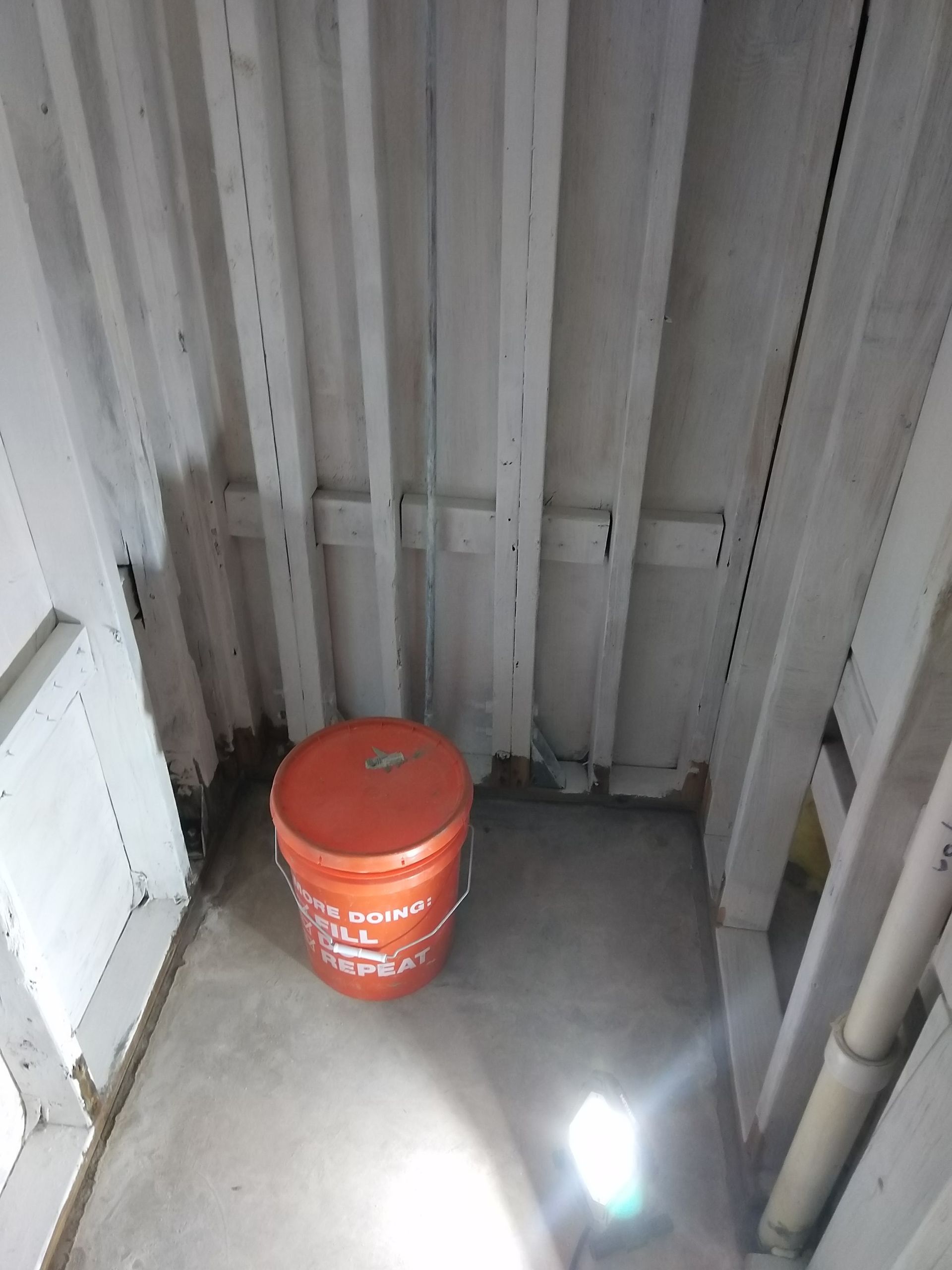 Orange bucket on a concrete floor in a narrow white-walled storage corner.