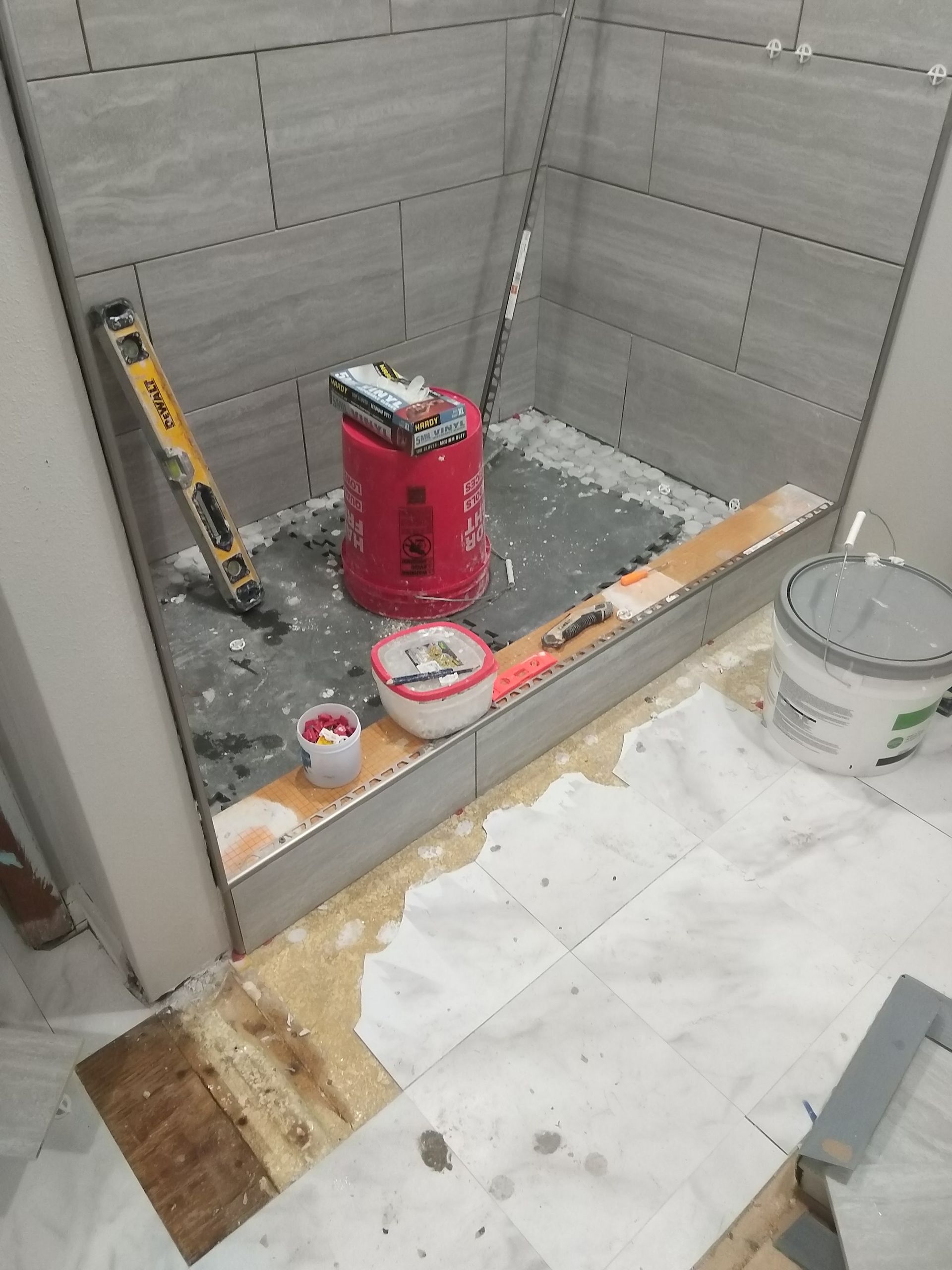 Construction area in a tiled shower stall with a red tool bucket, paint cans, and debris on the floor