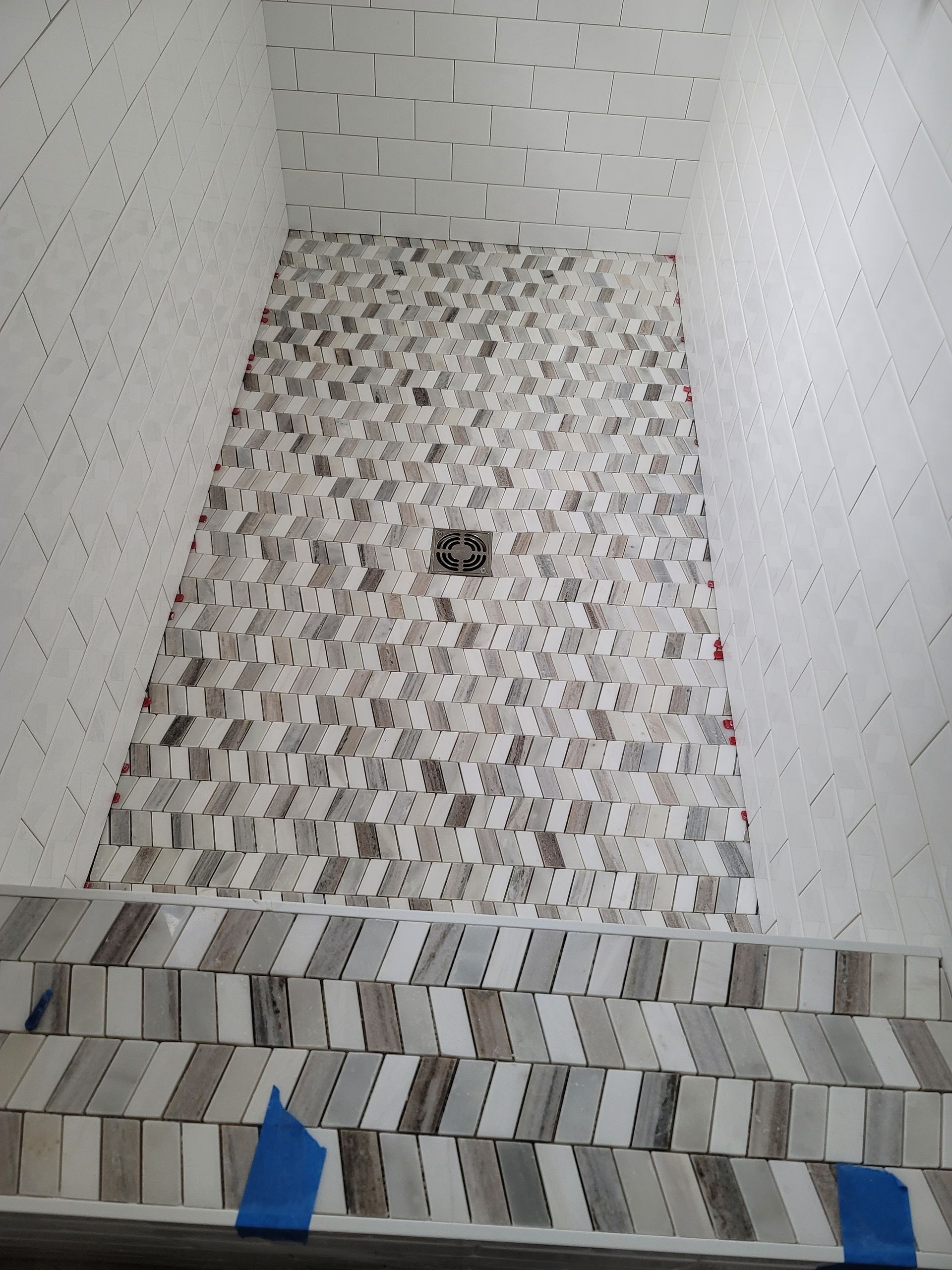 Tiled shower stall with a drain in the center and blue tape on the floor edge.