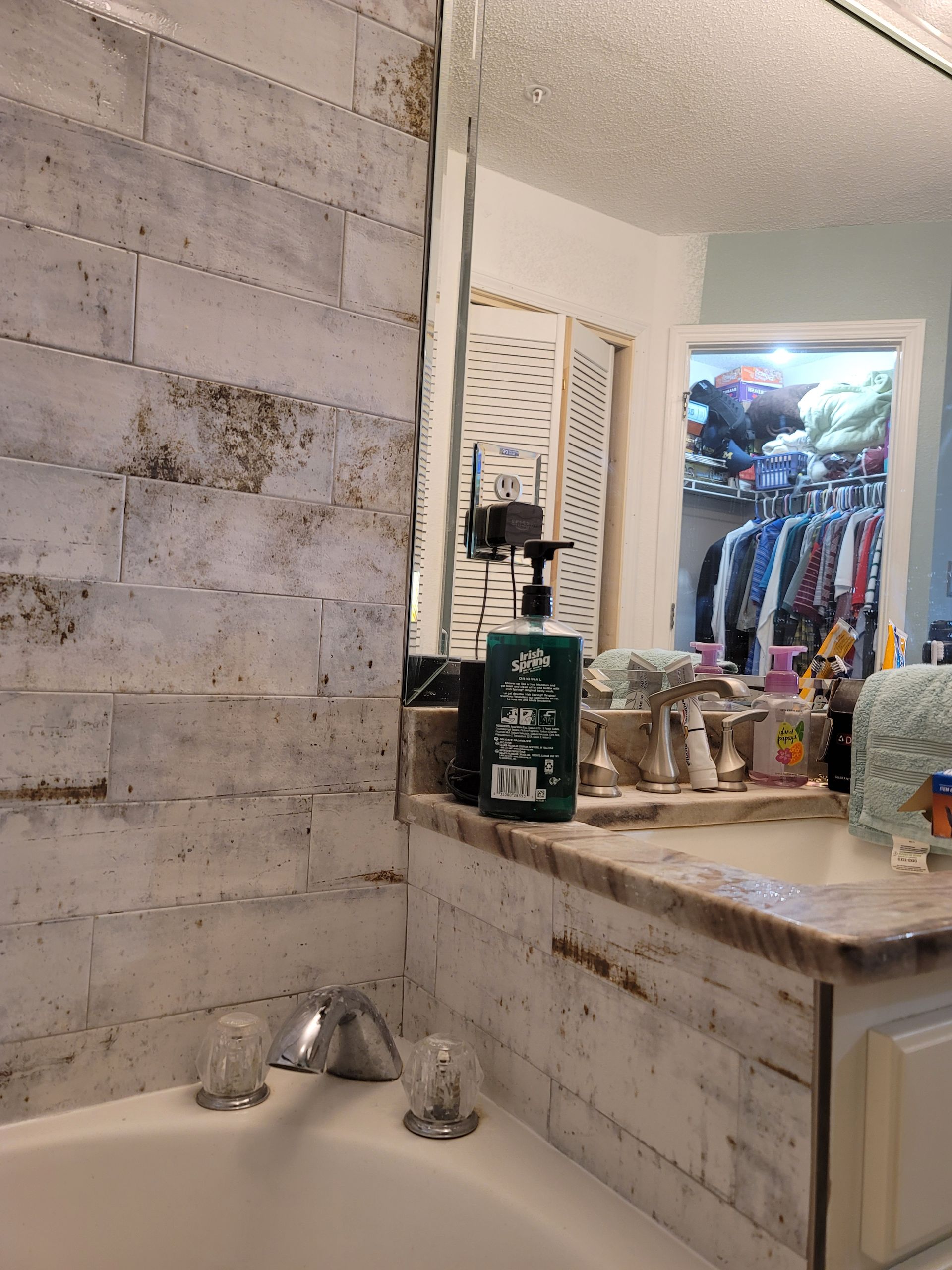 Bathroom vanity with whitewashed tile, sink, soap dispenser, and a view into a closet with hanging clothes.