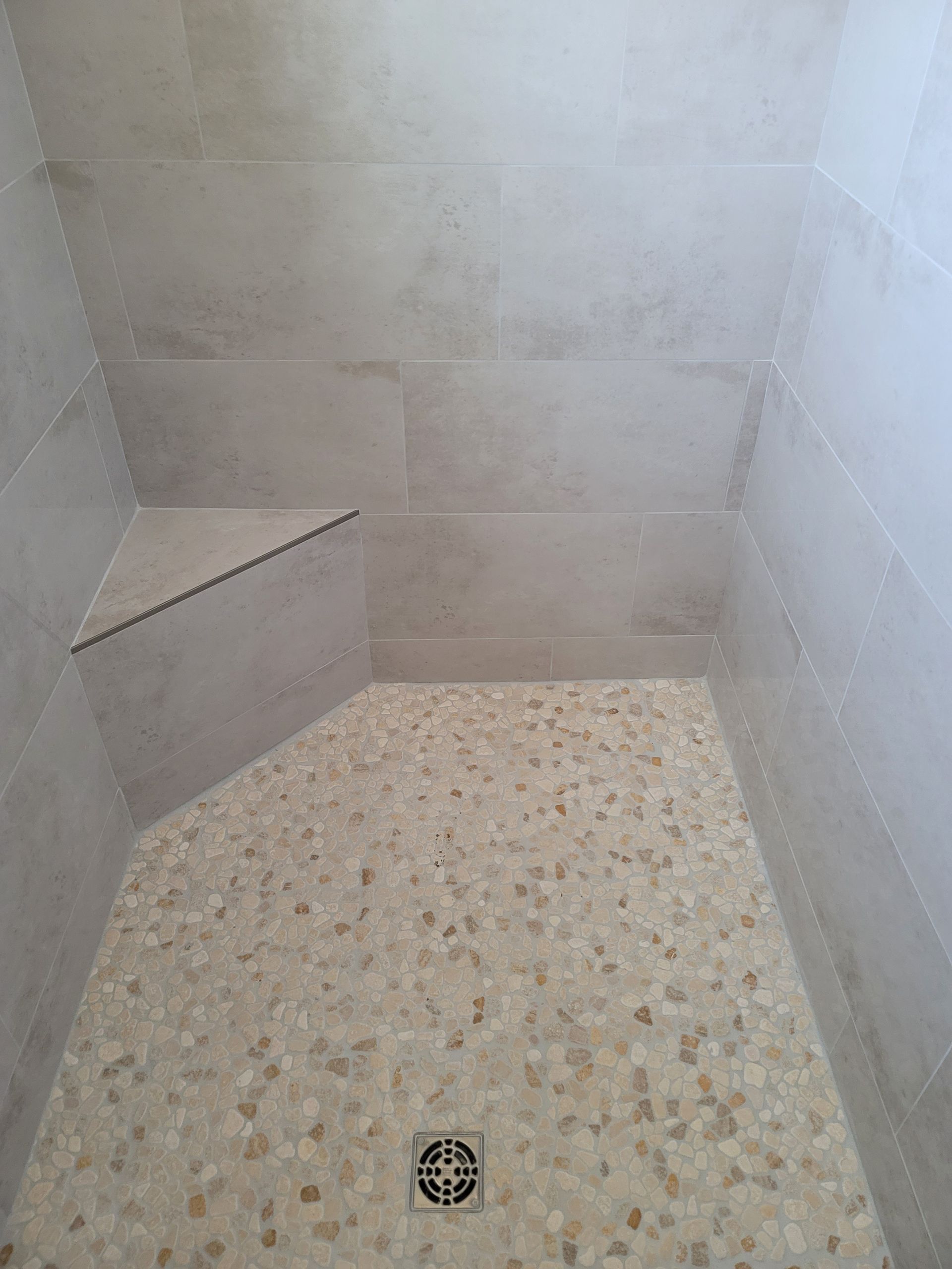 Small tiled shower stall with pebble floor, drain, and built-in corner shelf.