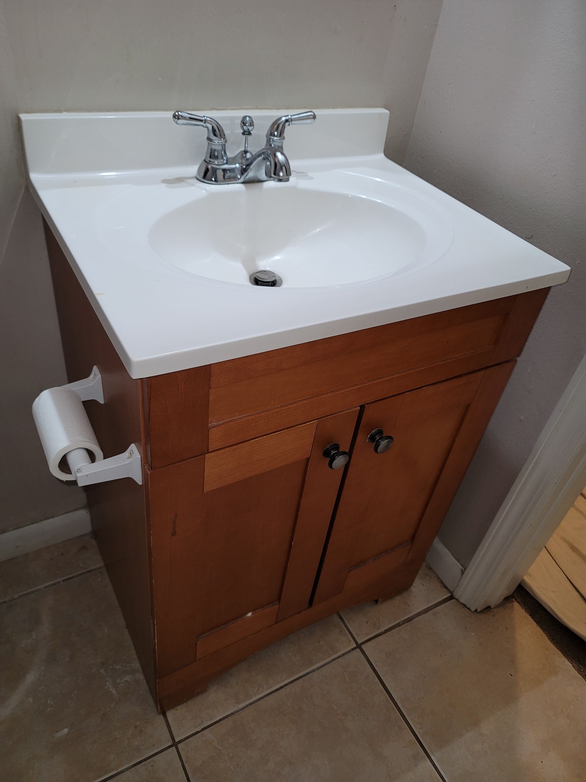 White bathroom sink vanity with wooden cabinet, chrome faucet, and toilet paper holder beside tiled floor.