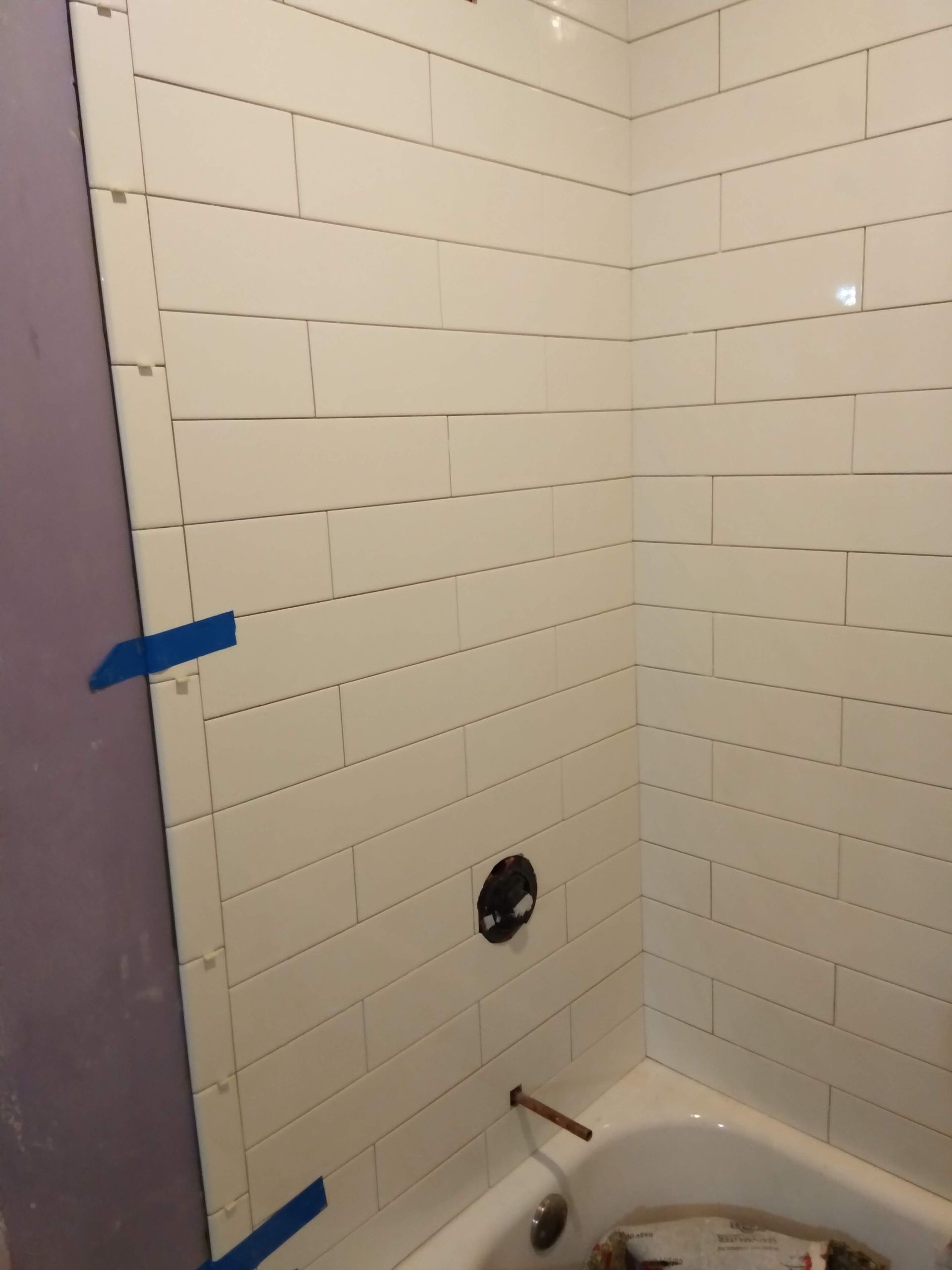 Partially tiled bathroom shower corner with a bathtub, exposed outlet, and blue painter’s tape on the wall
