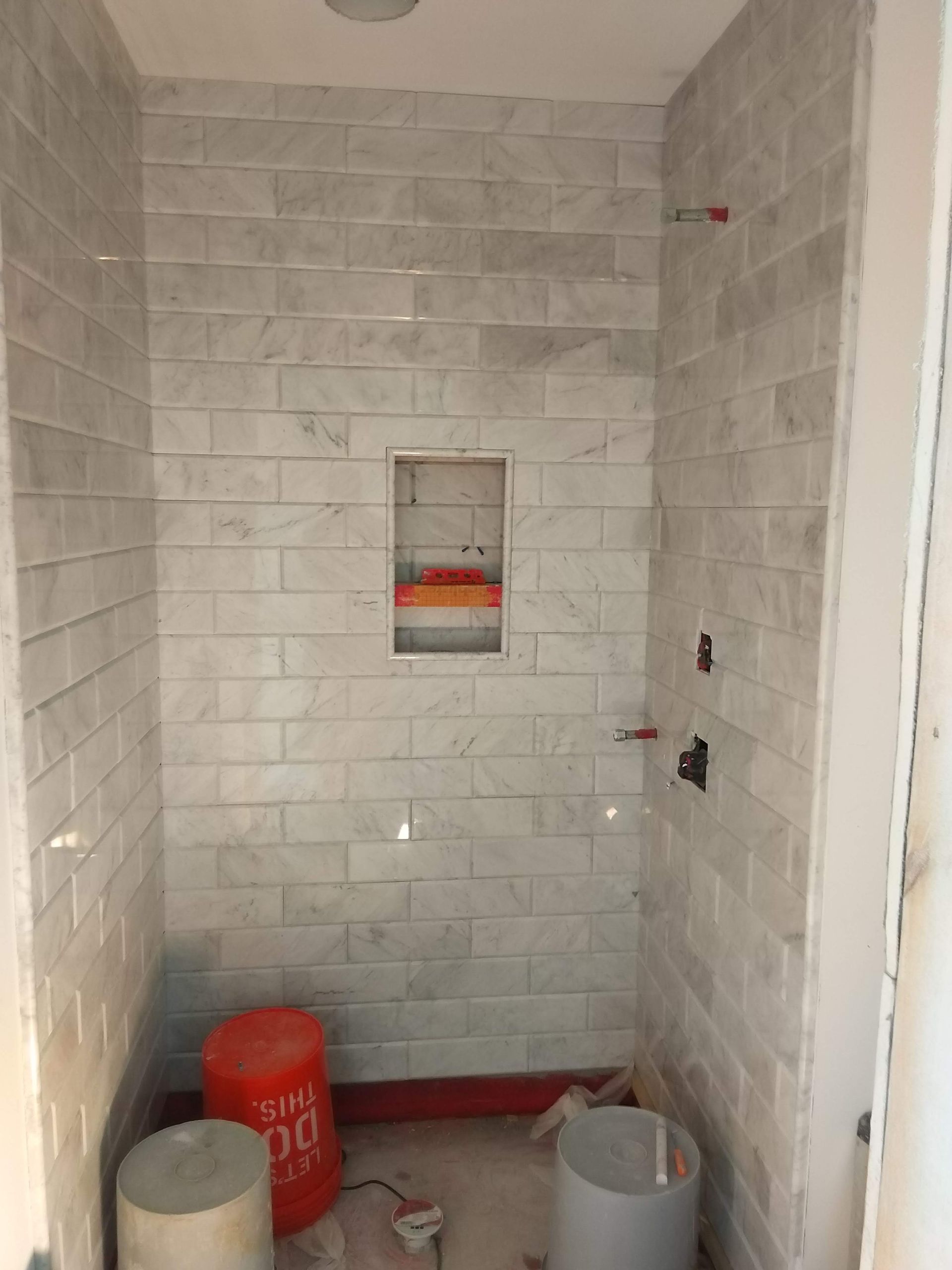 Small tiled shower stall under construction with exposed plumbing and buckets on the floor