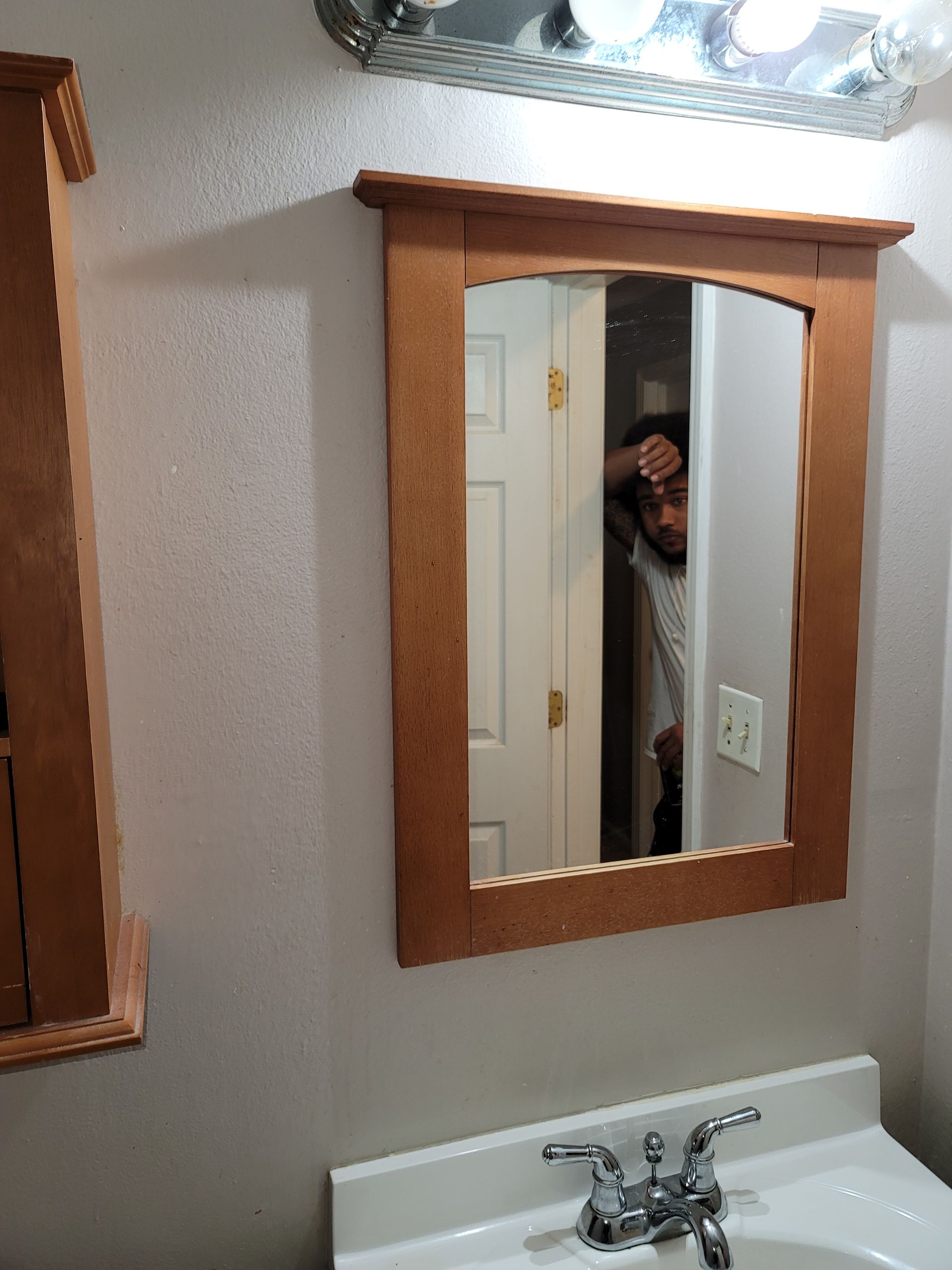 Bathroom mirror with a person peeking from a doorway, vanity light above a wooden-framed mirror.