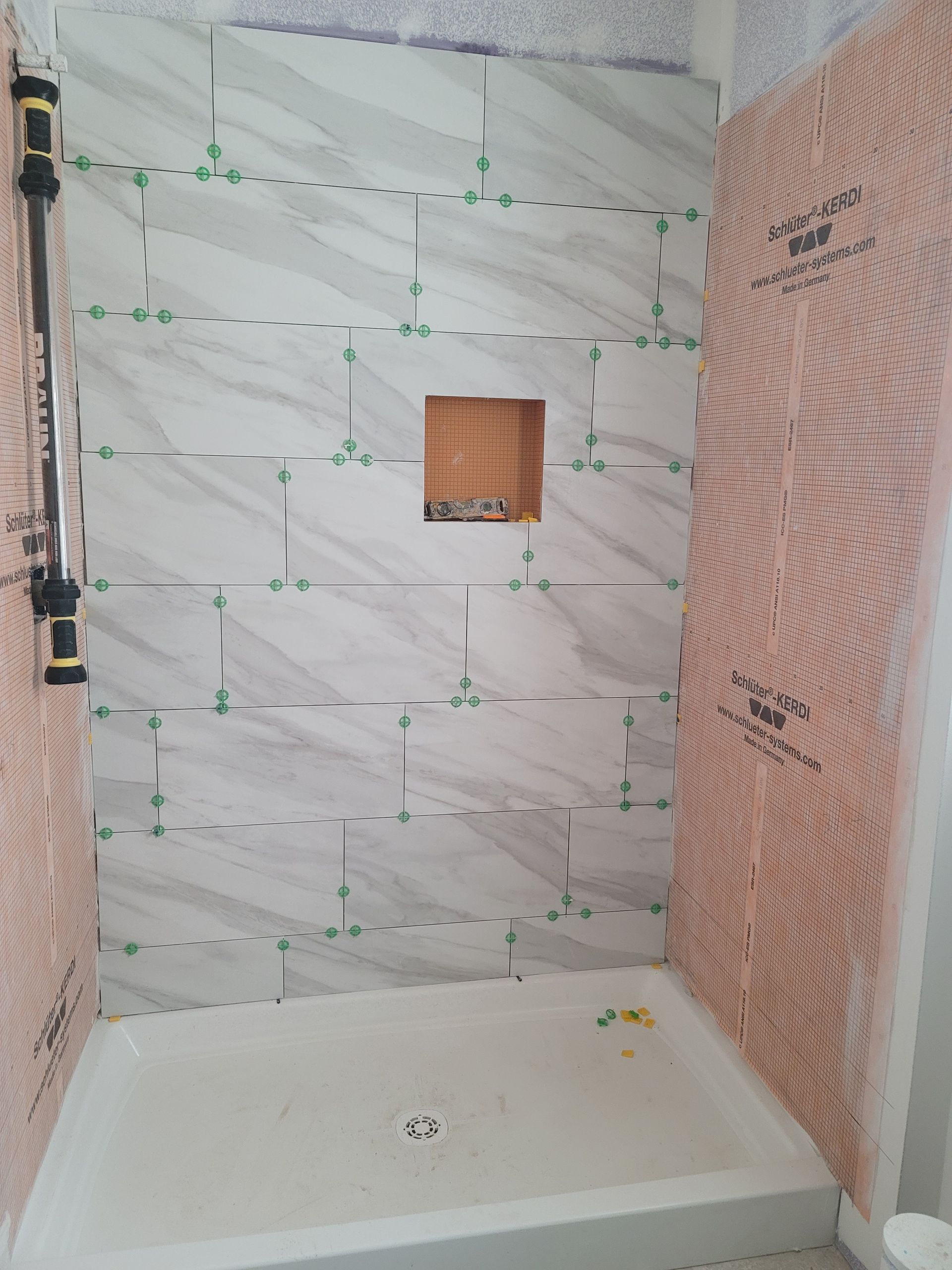 Partially tiled shower with a white tub, marble-look back wall, and exposed plumbing opening.