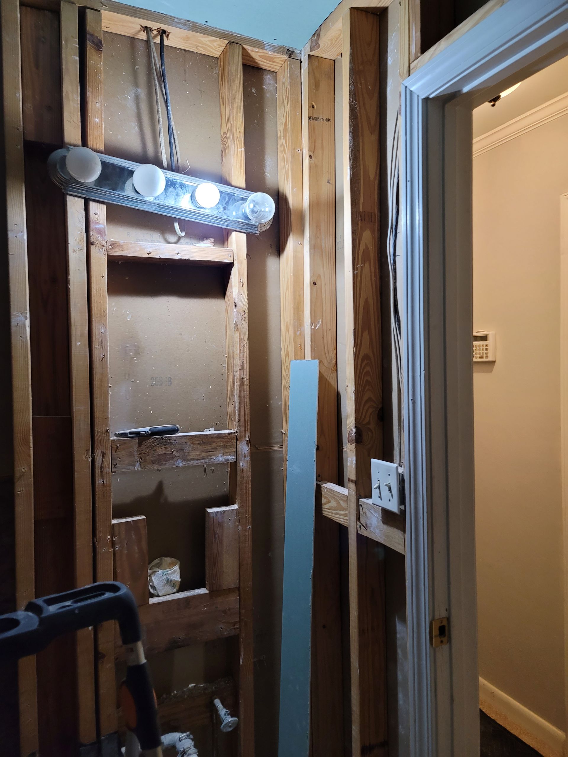 Bathroom wall under renovation with exposed studs, hanging light fixture, and open doorway
