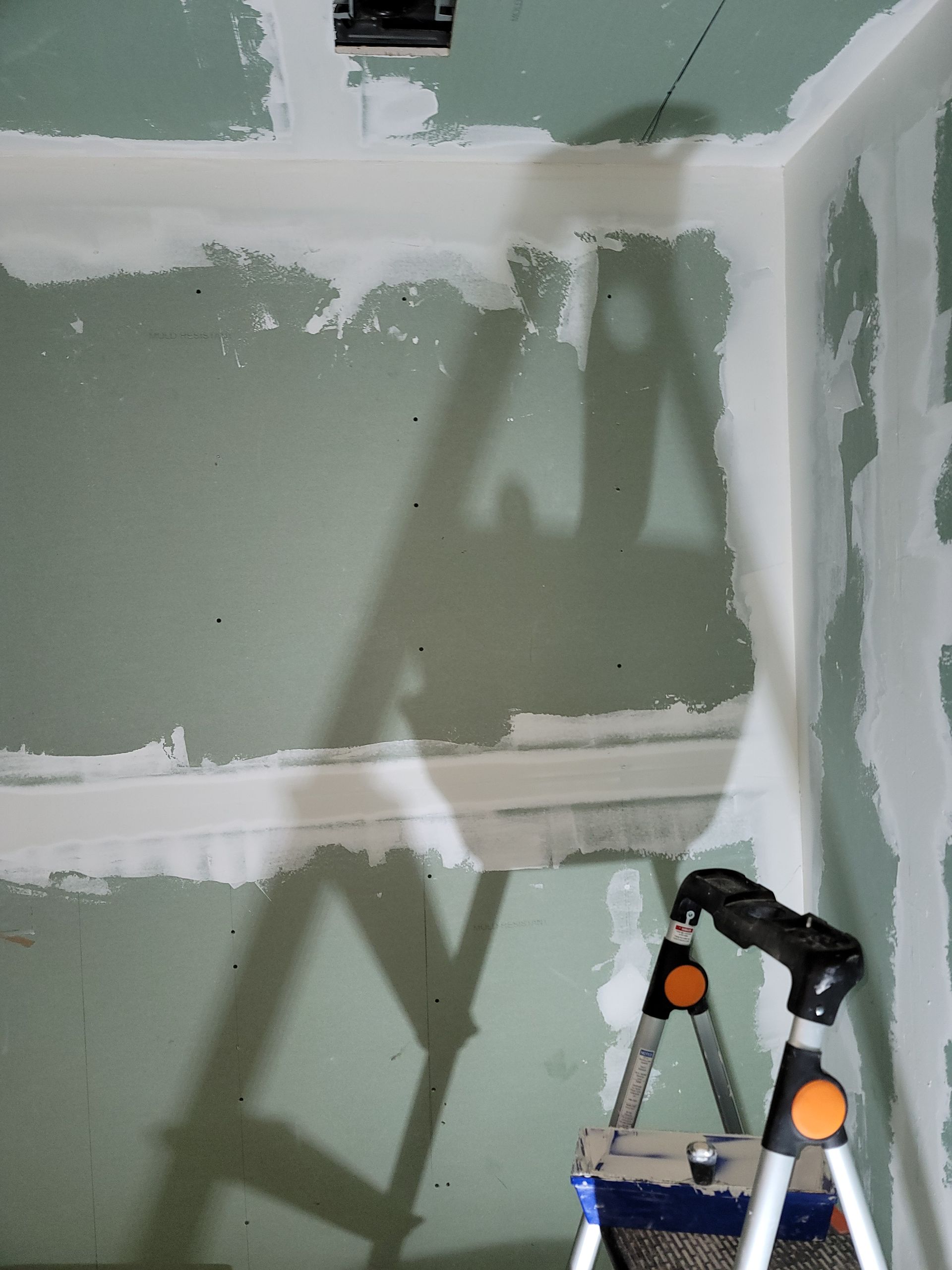 Unfinished drywall corner with white joint compound and a step ladder casting a shadow.