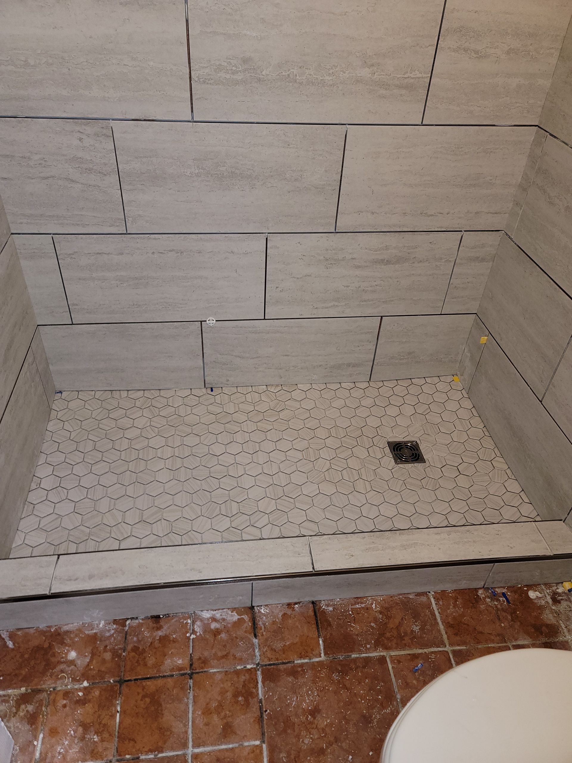 Tiled shower stall with pebble shower floor and a small drain, seen above terracotta floor tiles.