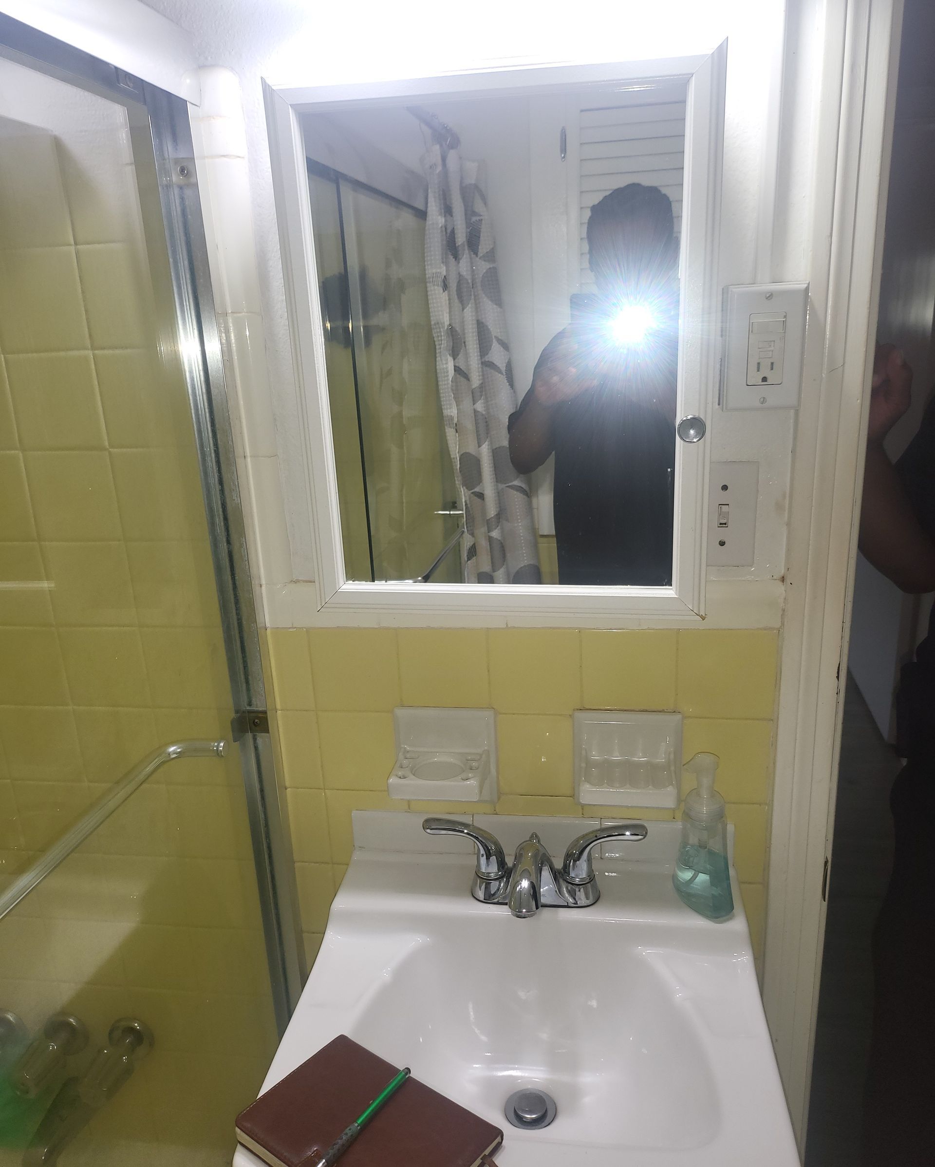 Bathroom sink and mirror with yellow tiled walls; person reflected taking a photo.