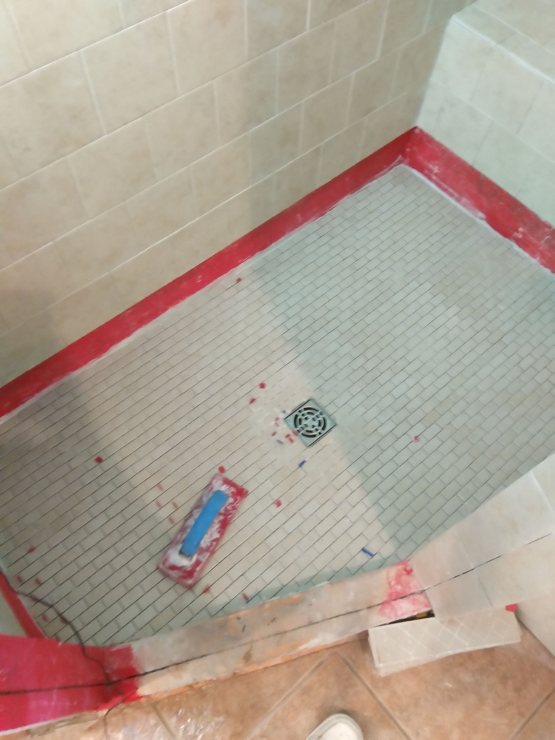 Small tiled shower stall with a floor drain, red caulk edges, and a tube of sealant on the floor