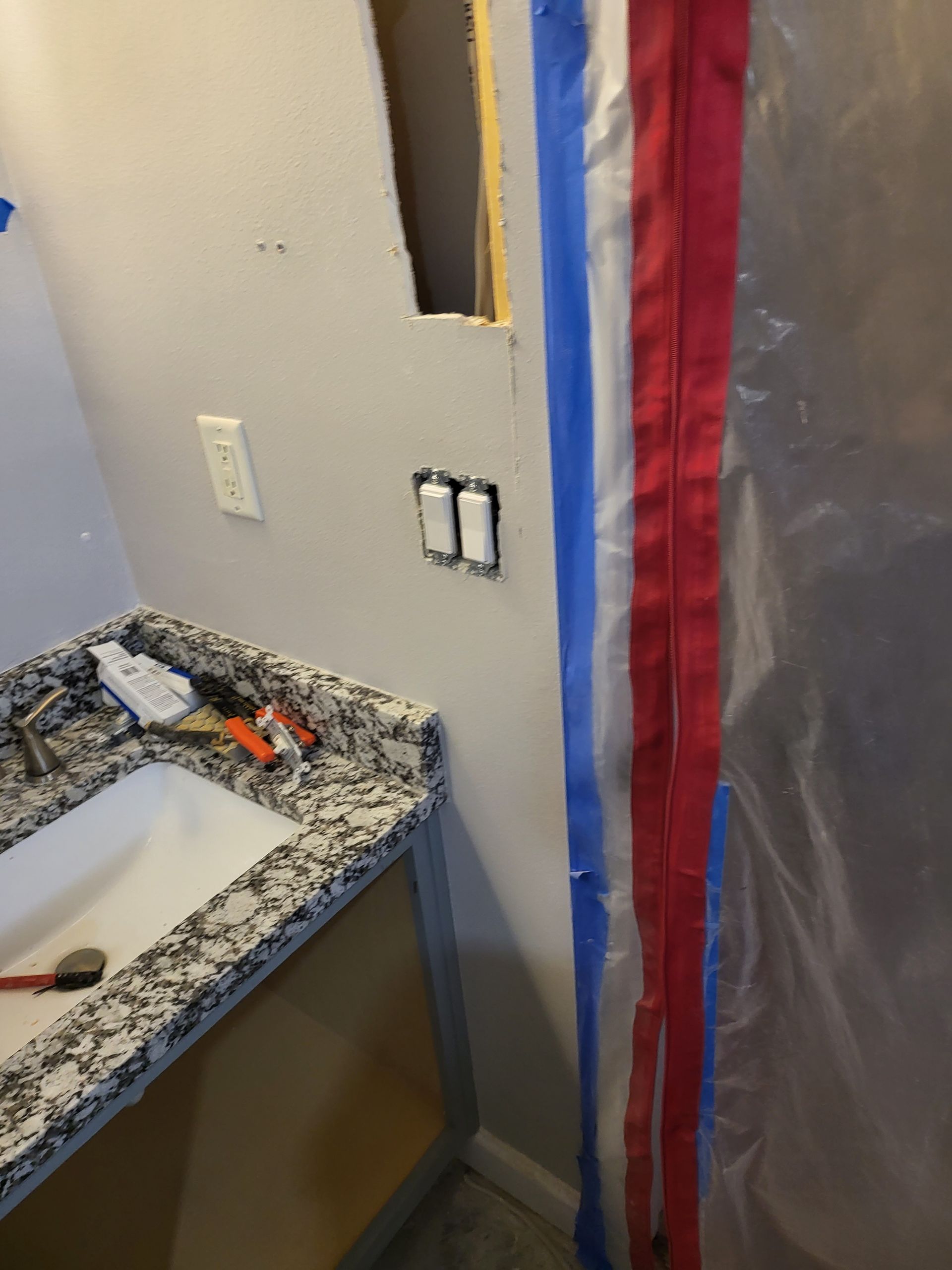Bathroom wall under renovation with exposed sink edge, outlets, and plastic sheeting by a red-trimmed doorway