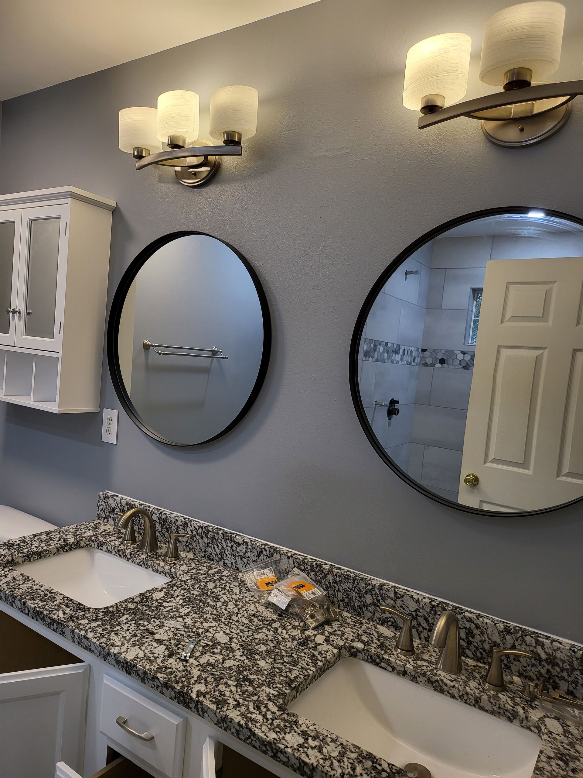 Bathroom vanity with two oval mirrors, granite countertop, sinks, and wall lights reflected in the mirror.