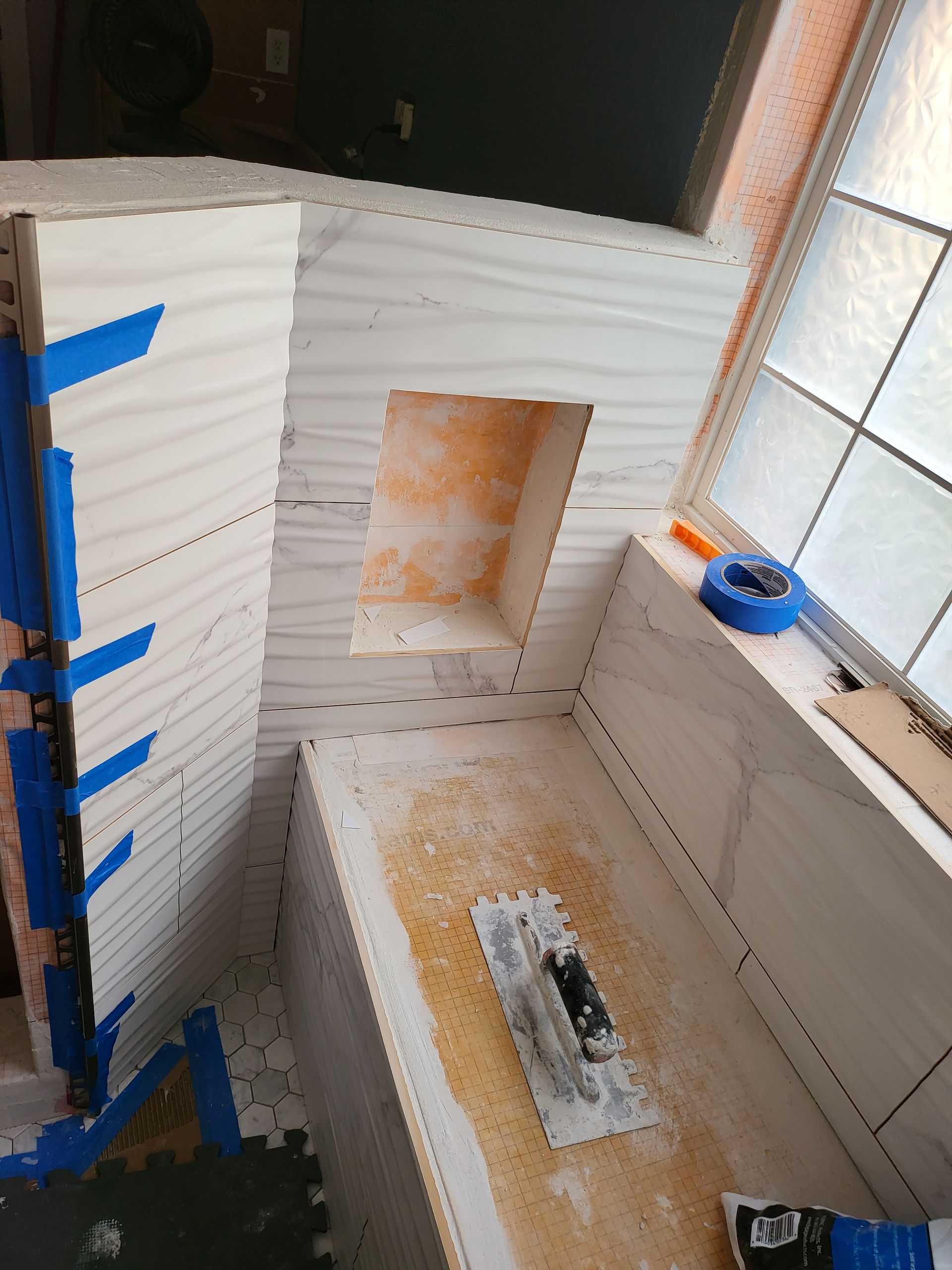 Small bathroom under renovation with tiled walls, a wall niche, blue tape, and construction debris on the floor.
