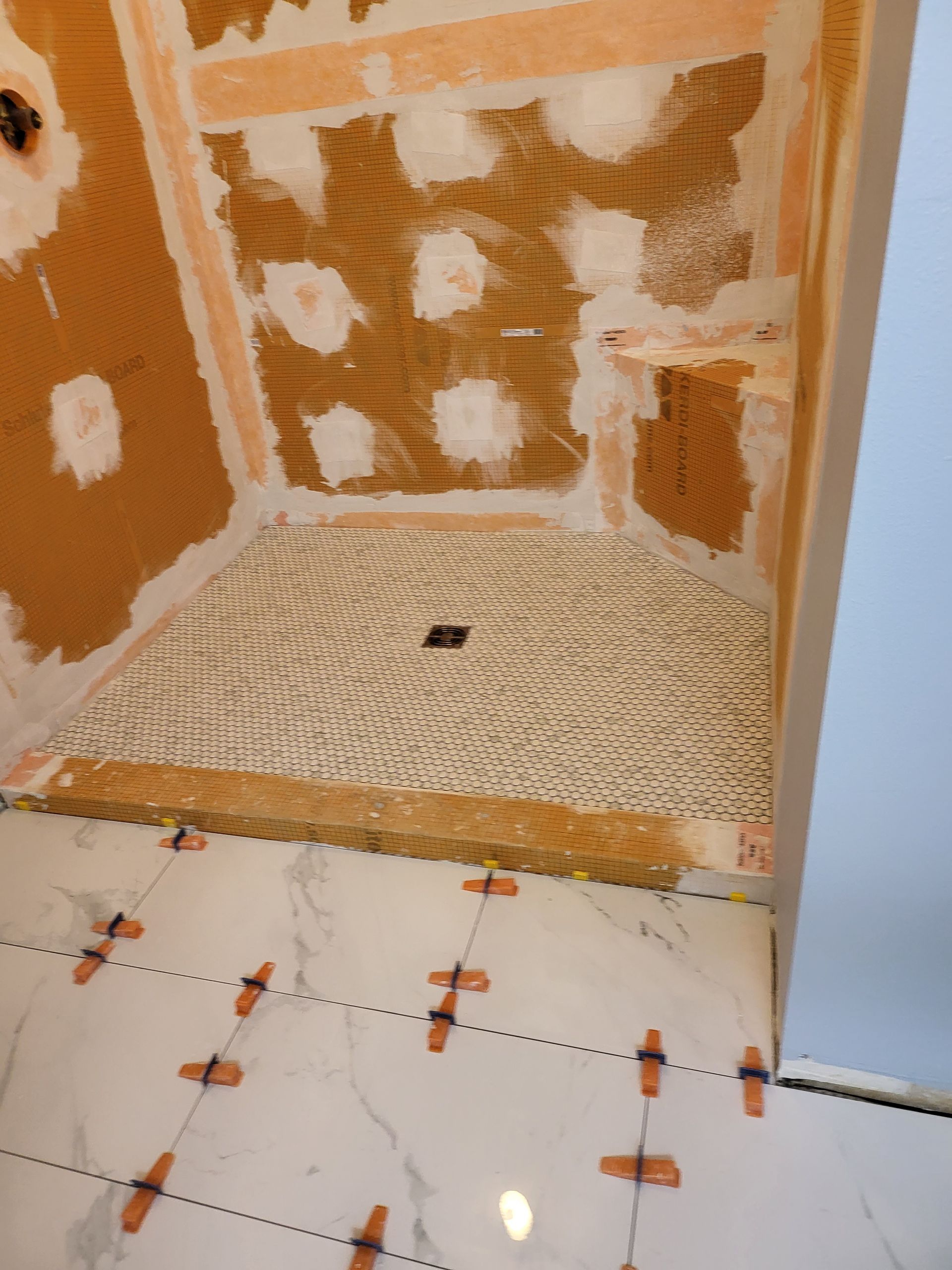 Tiled shower corner with orange waterproofing, unfinished walls, and a partially tiled floor with spacers