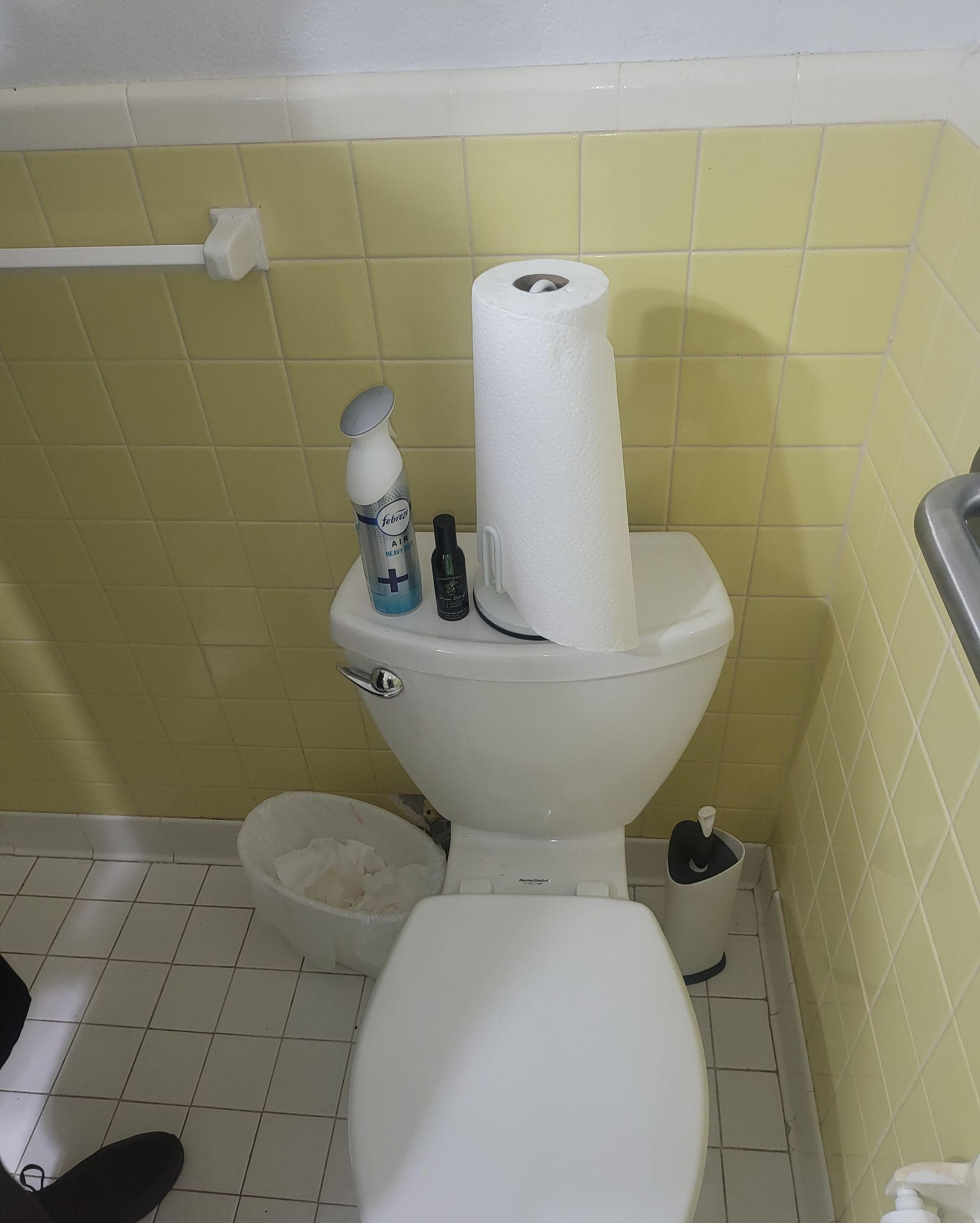 Small bathroom with a toilet tank holding a paper towel roll, cleaning bottles, and pale yellow tile walls.