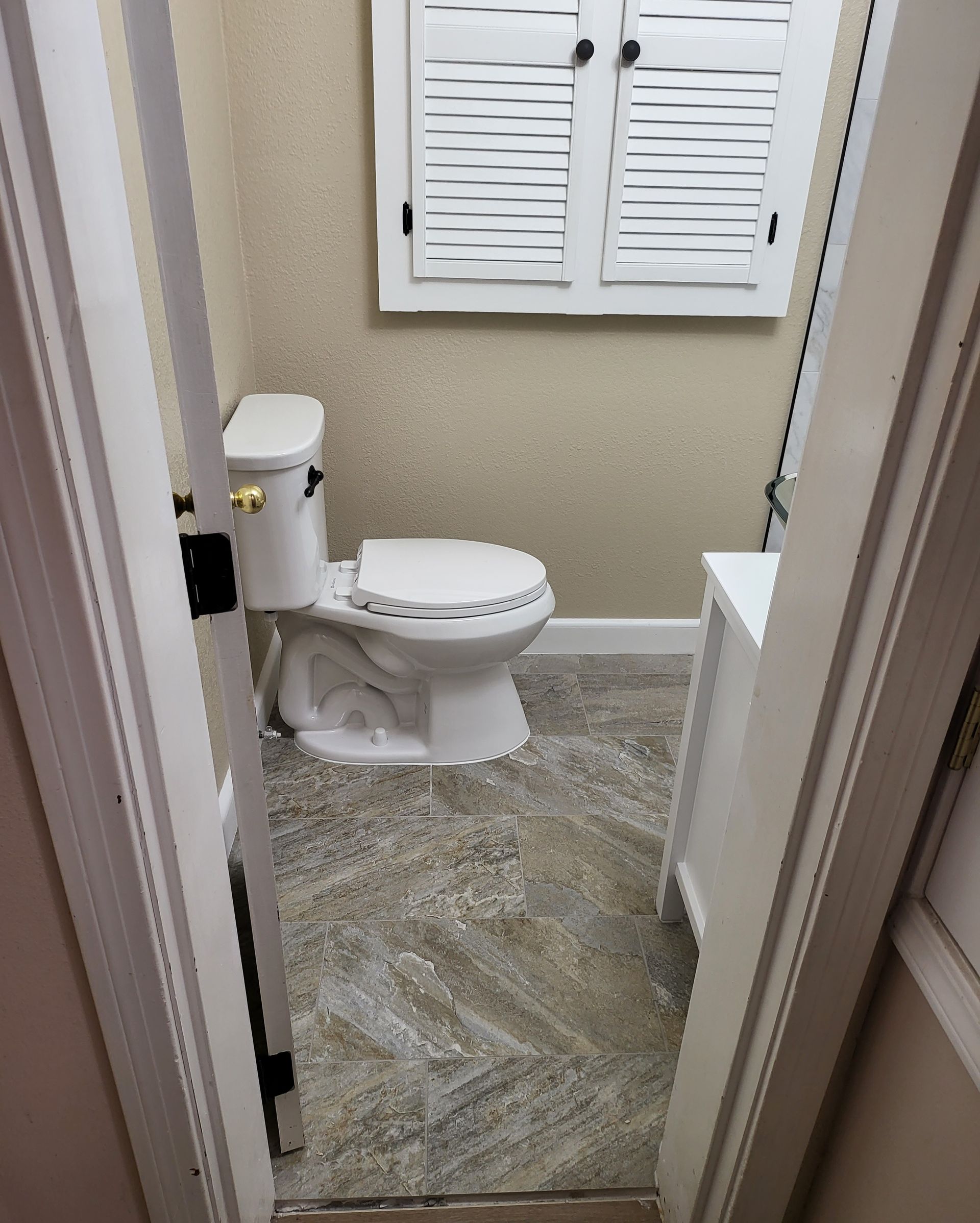 Small bathroom with a toilet, gray tile floor, white shuttered cabinet, and beige walls.