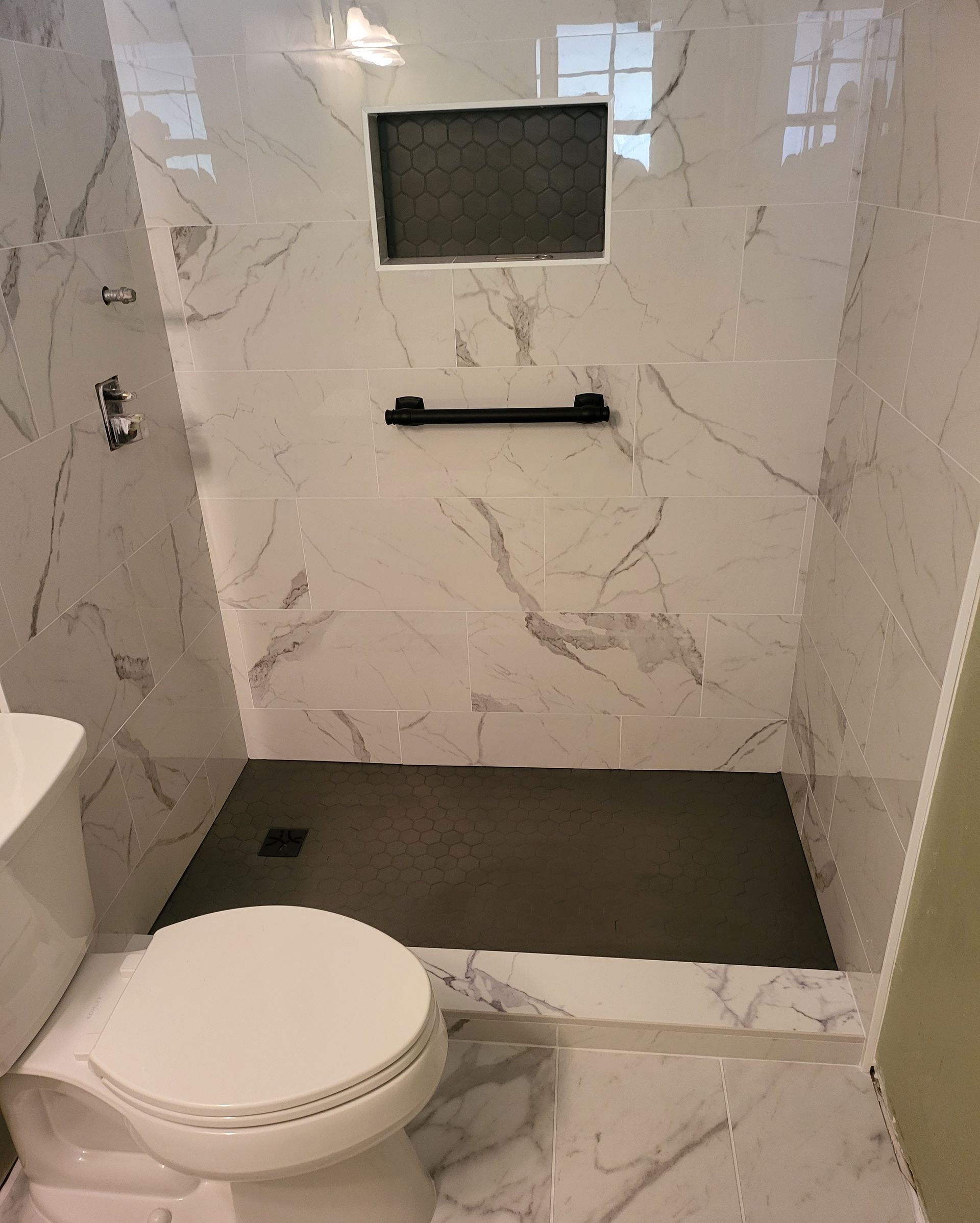Small modern shower with white marble walls, black shelf, and gray tile floor beside a toilet.