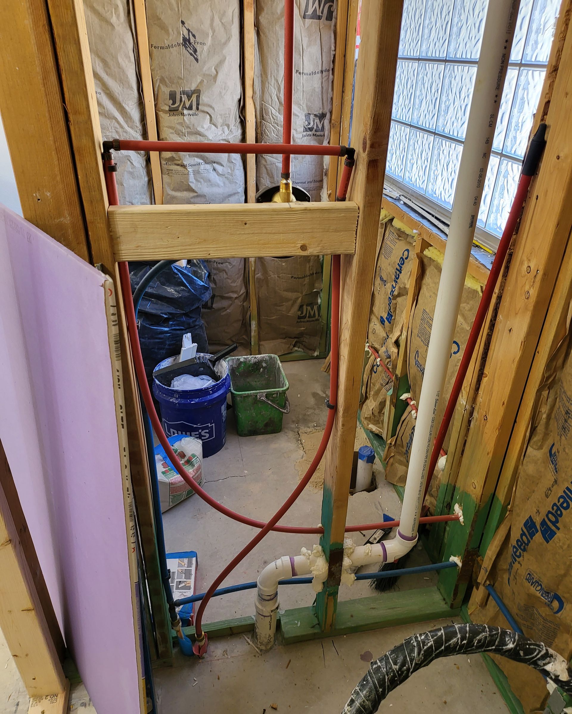Construction site with exposed wall framing, pipes, and buckets in a narrow interior space