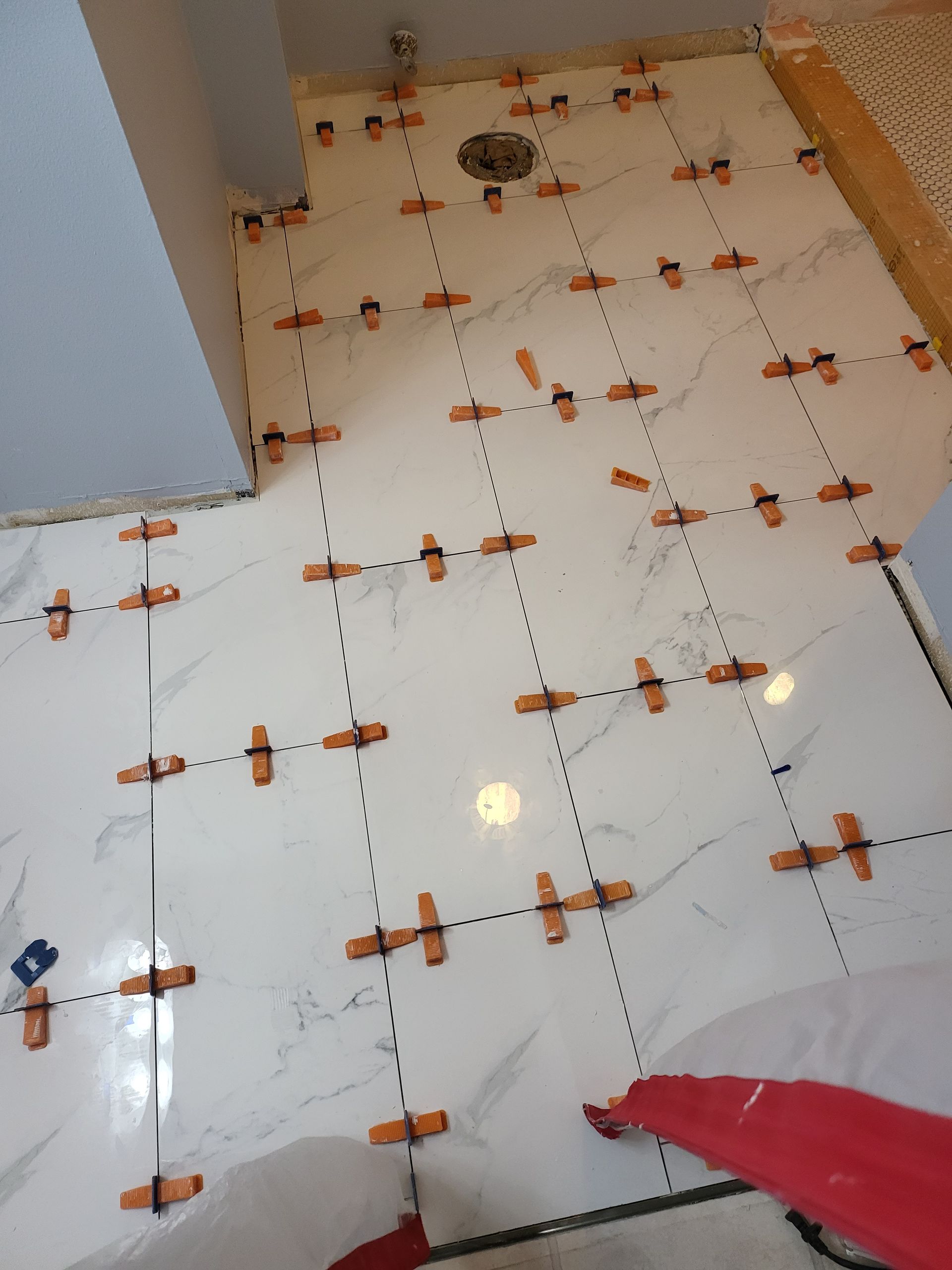 Tiled floor under renovation with orange leveling clips and spacers, plus a drain opening near the wall.