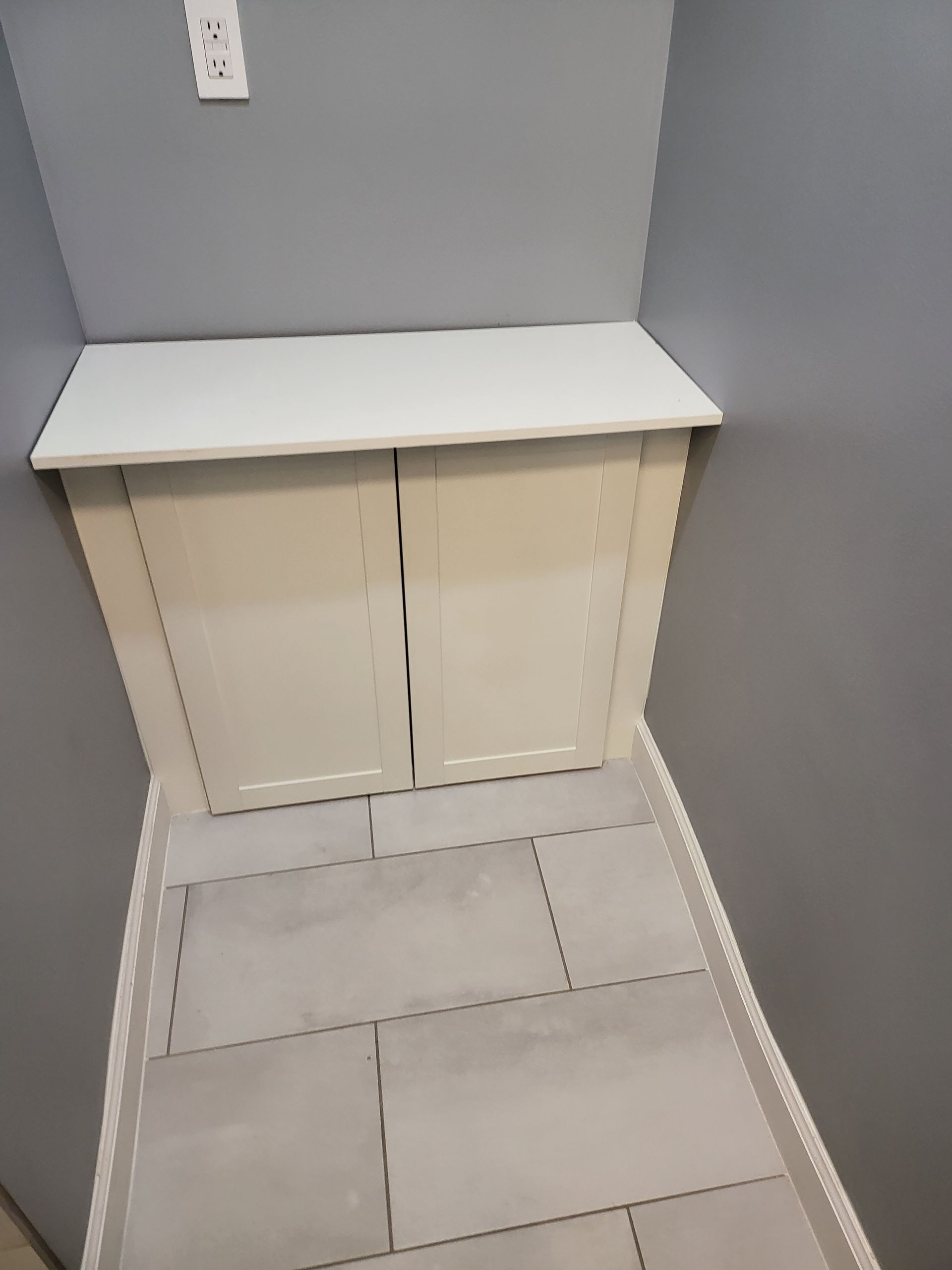 Small white cabinet in a gray corner with a countertop and tiled floor.