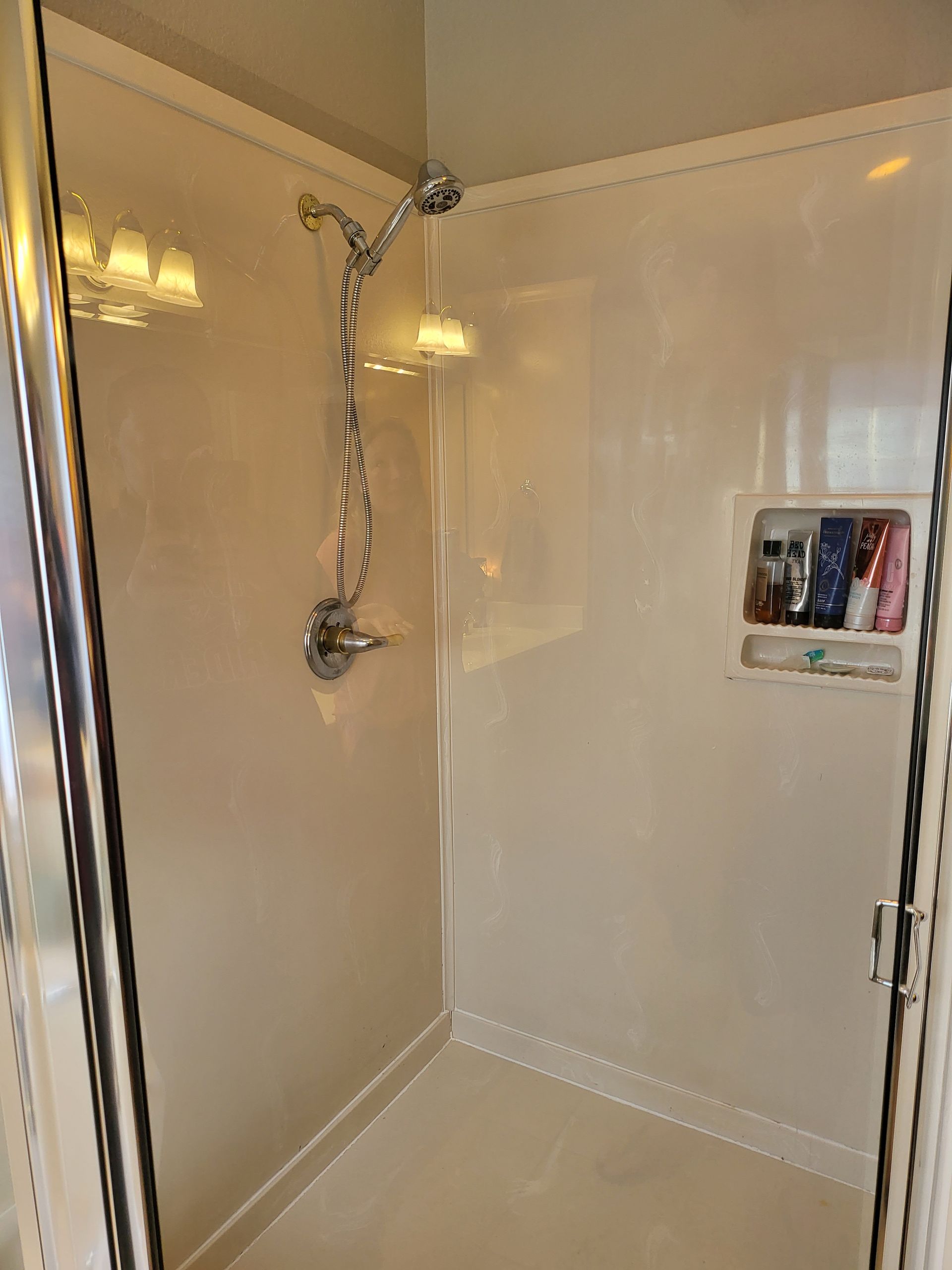 Cream-colored shower stall with chrome handheld shower and built-in wall niche with toiletries.