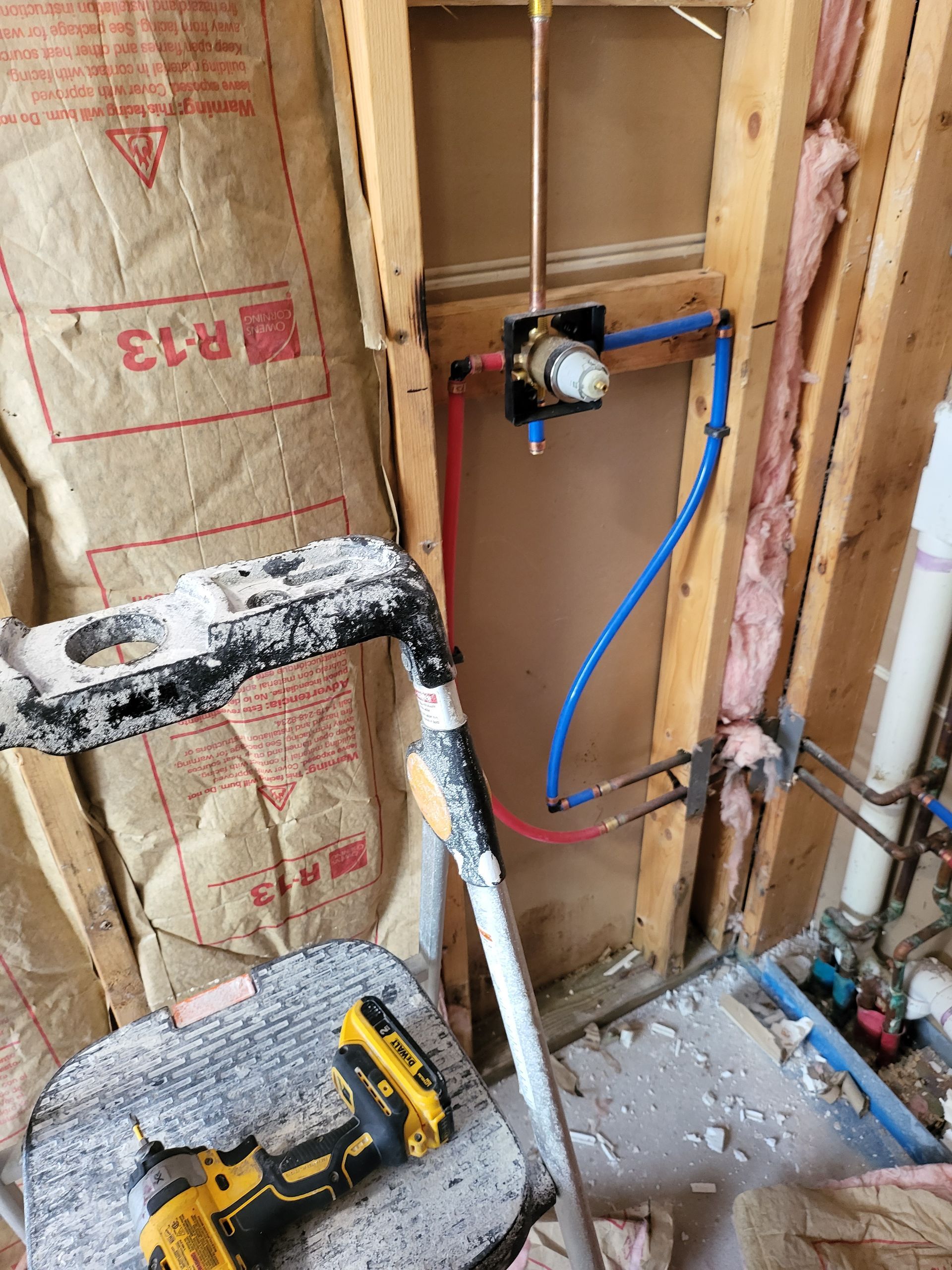 Partially built wall with plumbing fixtures, insulation, a drill on the floor, and a blue hose attached to a valve.