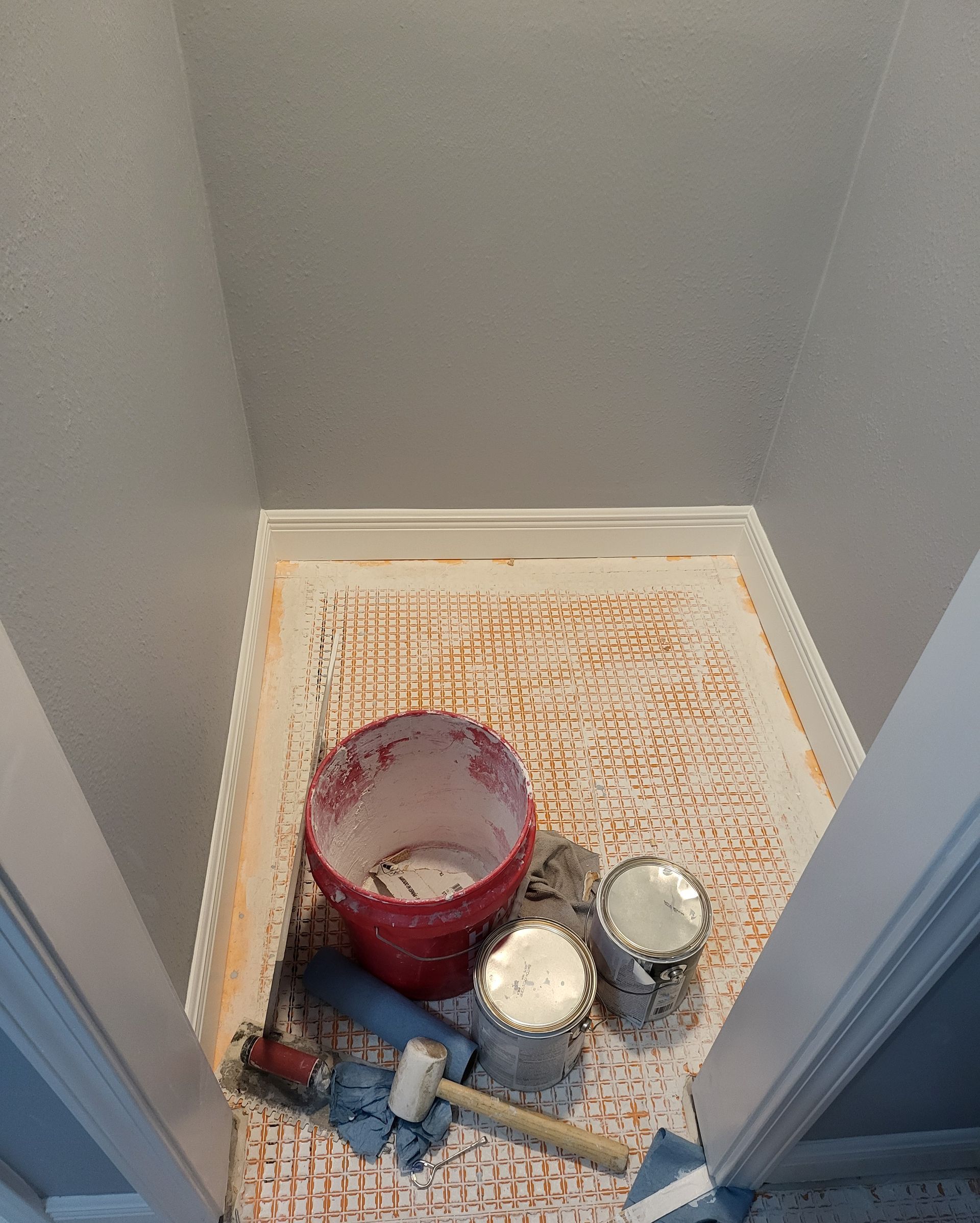 Paint cans and a brush on a tarp in a small room, with a red bucket and paint trays.