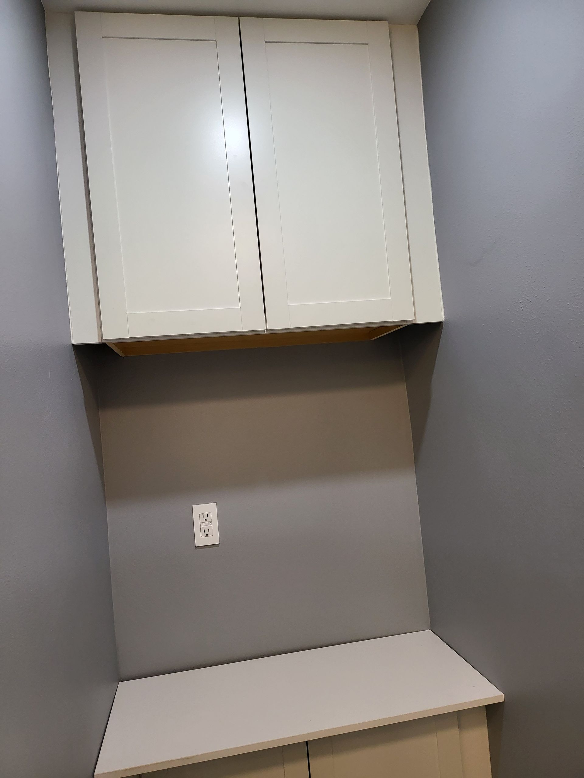 Small gray alcove with white upper cabinets, a wall outlet, and a light-colored countertop.