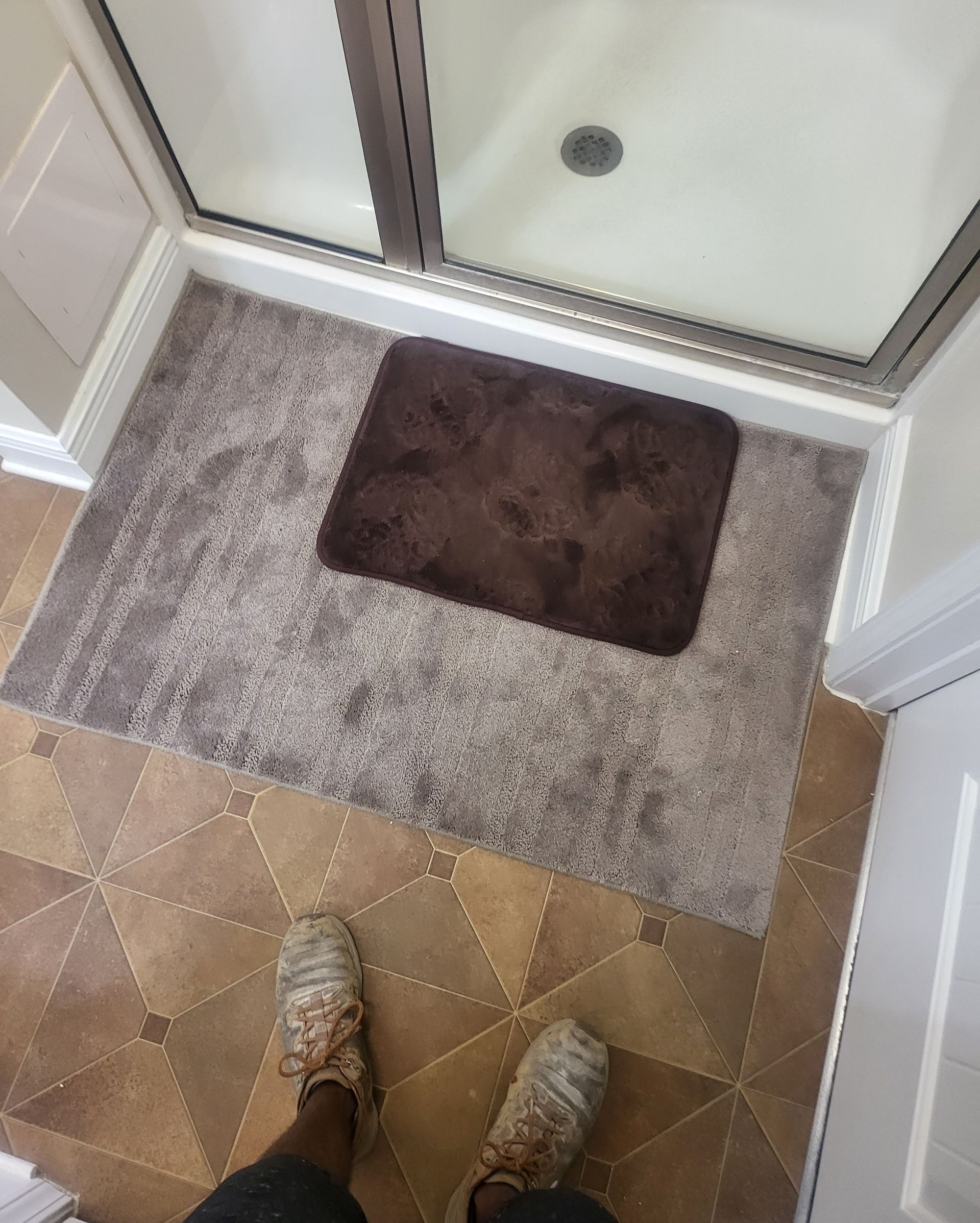 Shoes and feet on tan tile facing a shower entrance with a gray bath mat and brown rug