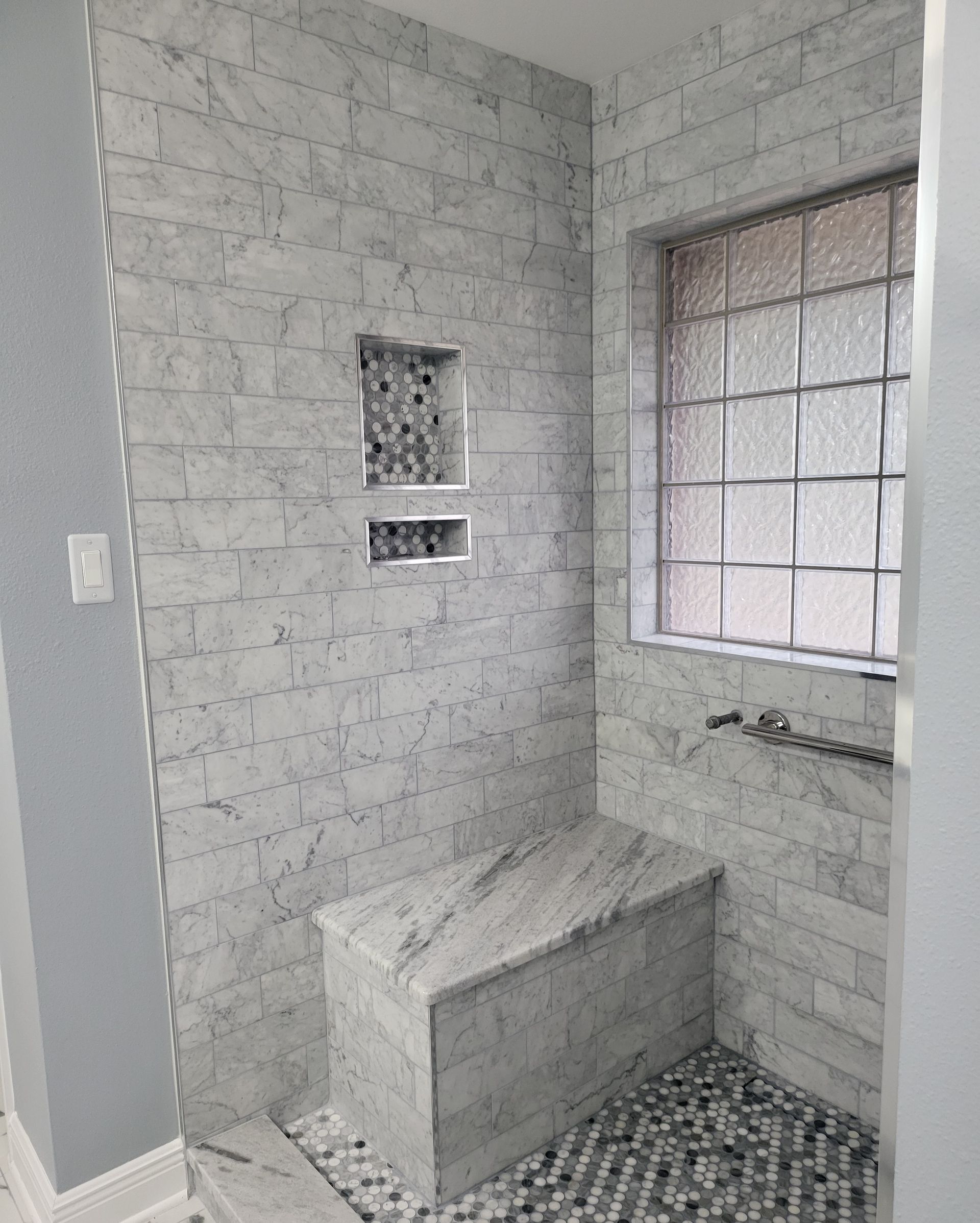 Small tiled shower with a built-in bench, window, and tiled floor in a gray bathroom.