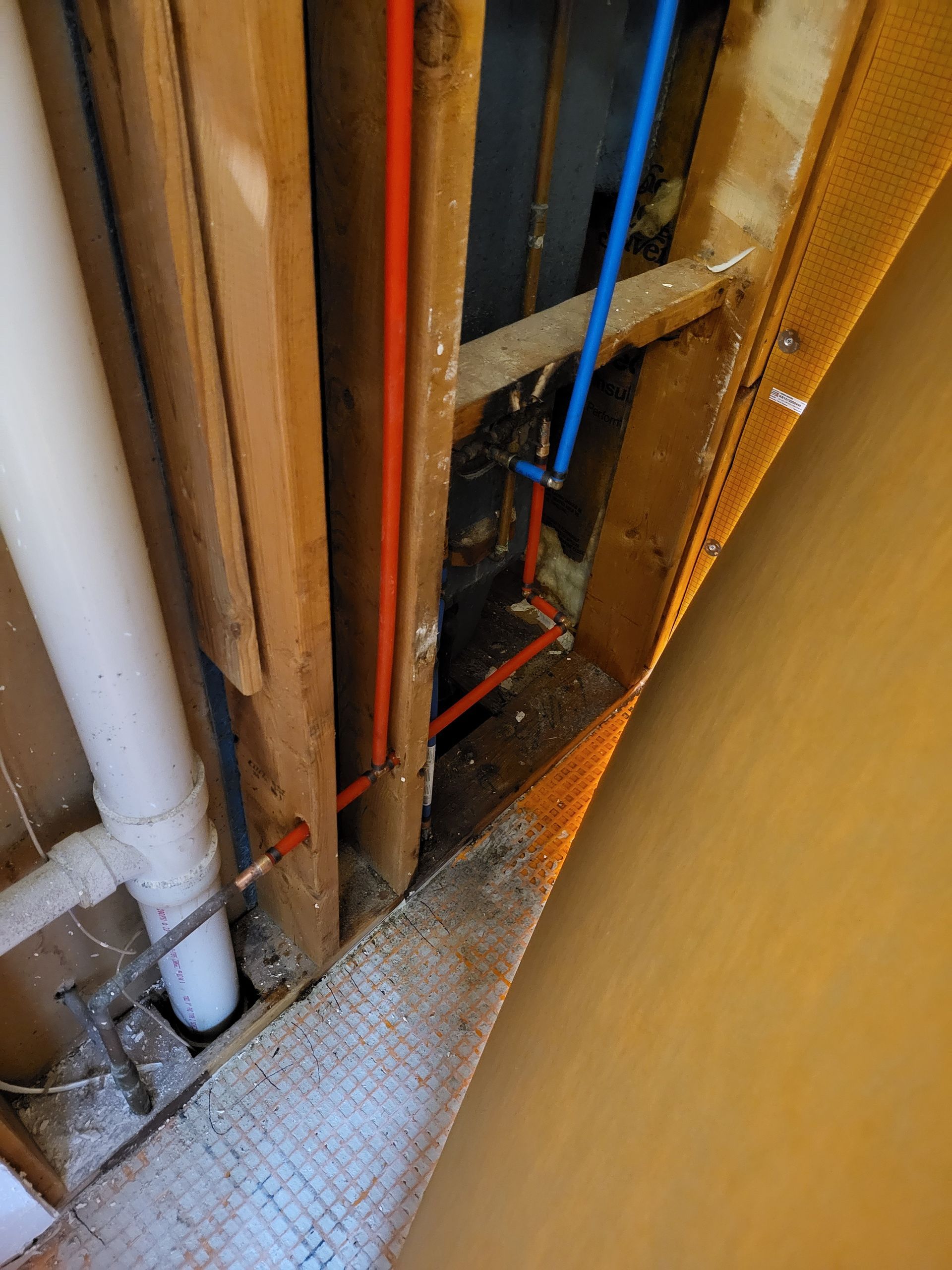 Narrow utility space with exposed pipes, wires, and blue plumbing lines beside a yellow wall and floor grate