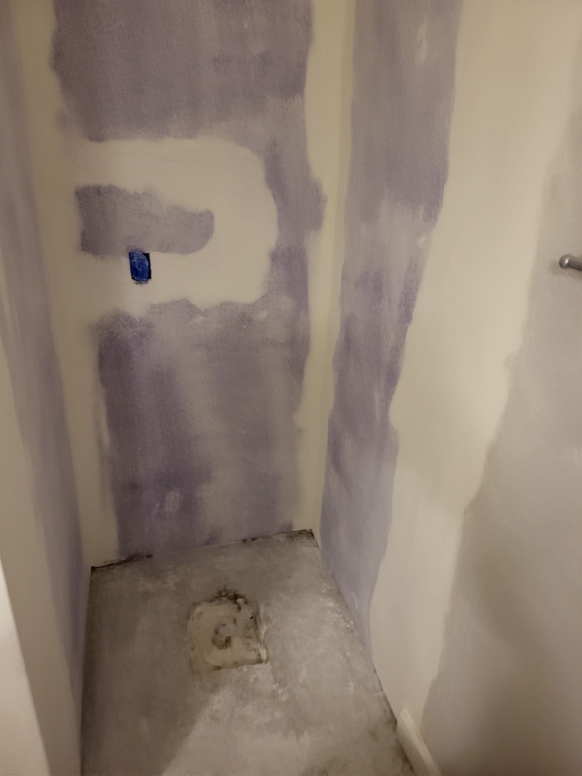 Small unfinished bathroom corner with purple drywall patches, exposed toilet plumbing, and a floor drain opening.
