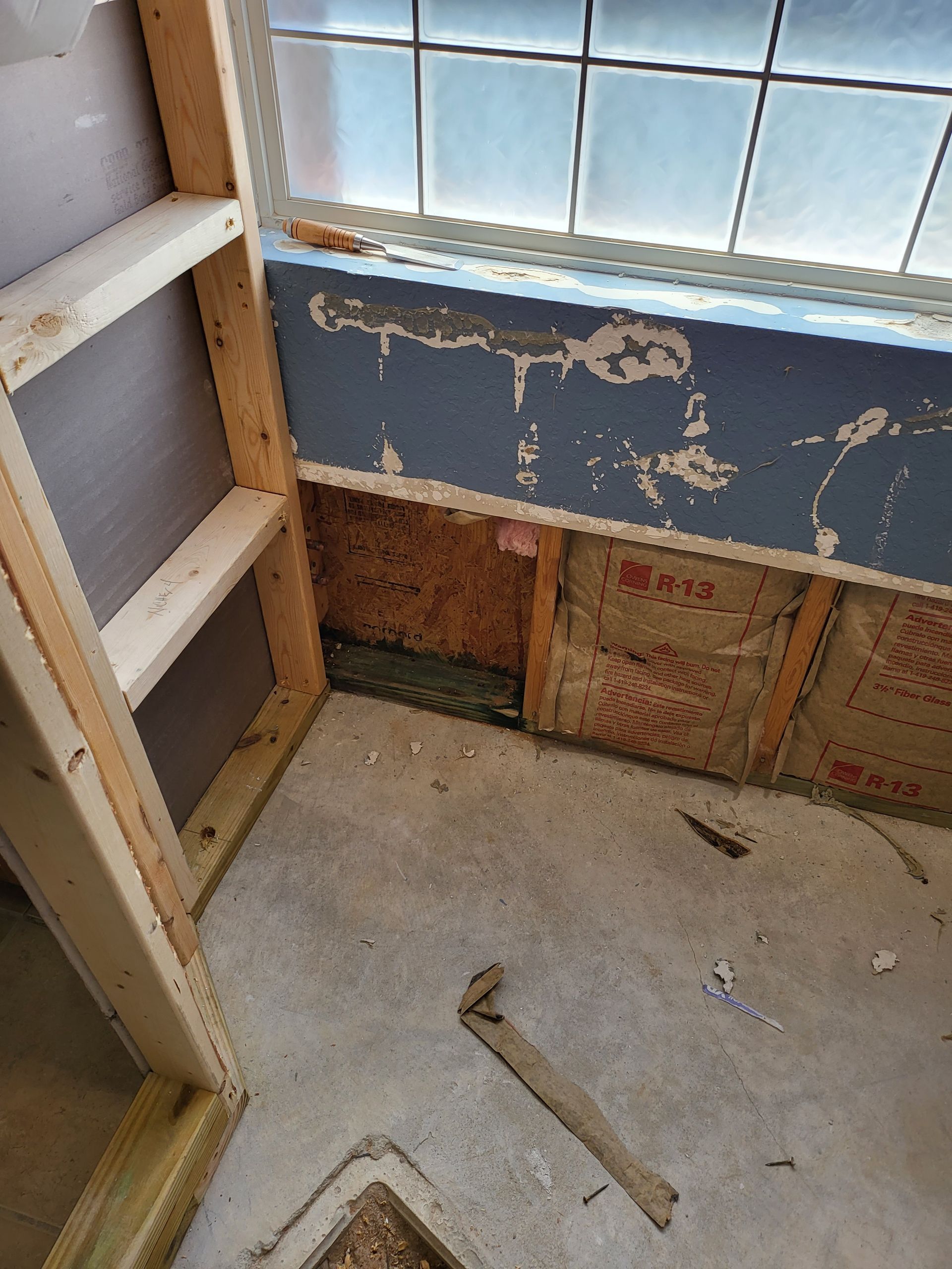 Partially stripped room wall with window, exposed insulation, ladder, and hammer on bare floor