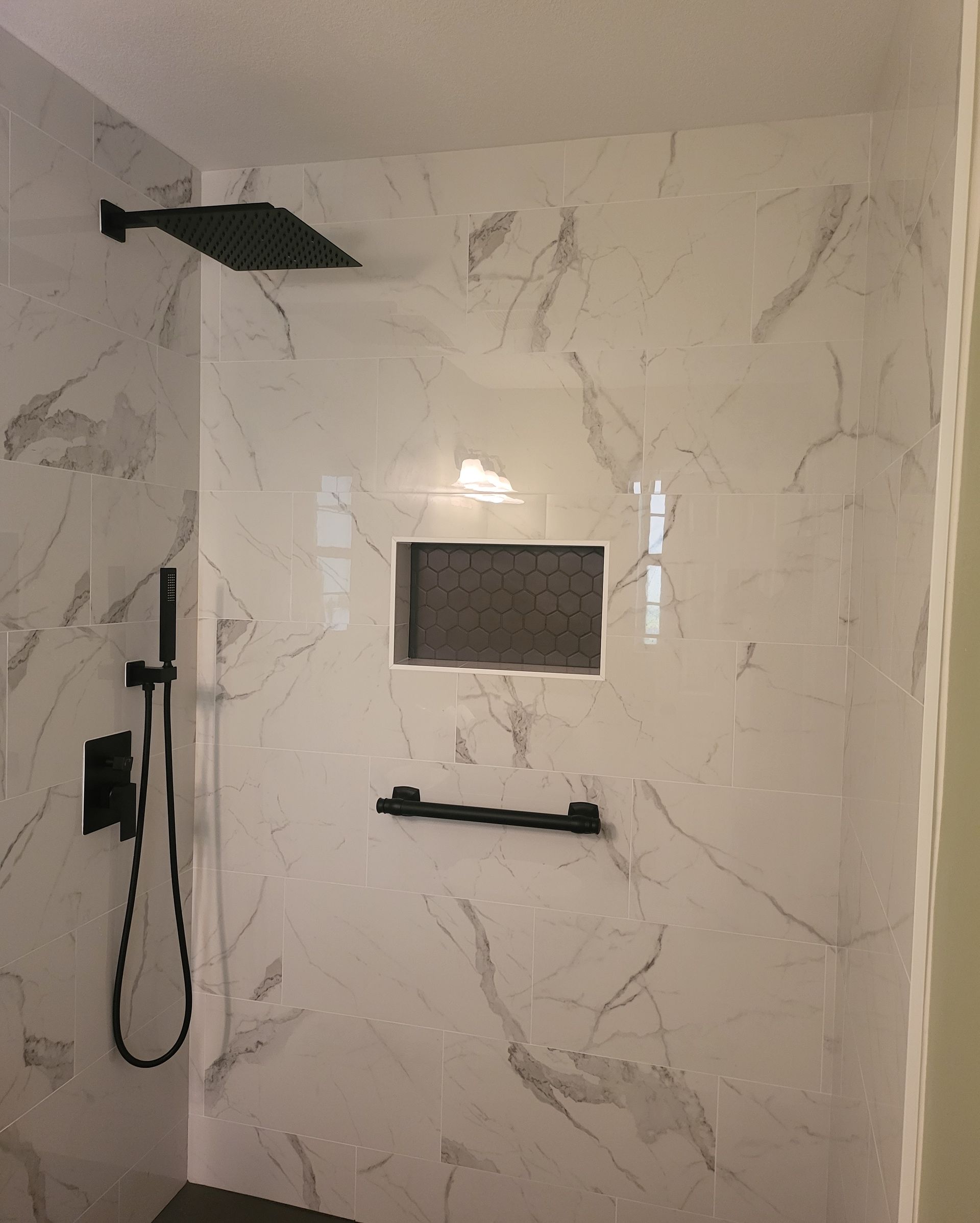 Modern walk-in shower with marble-pattern tile, black fixtures, showerhead, and a small recessed niche.