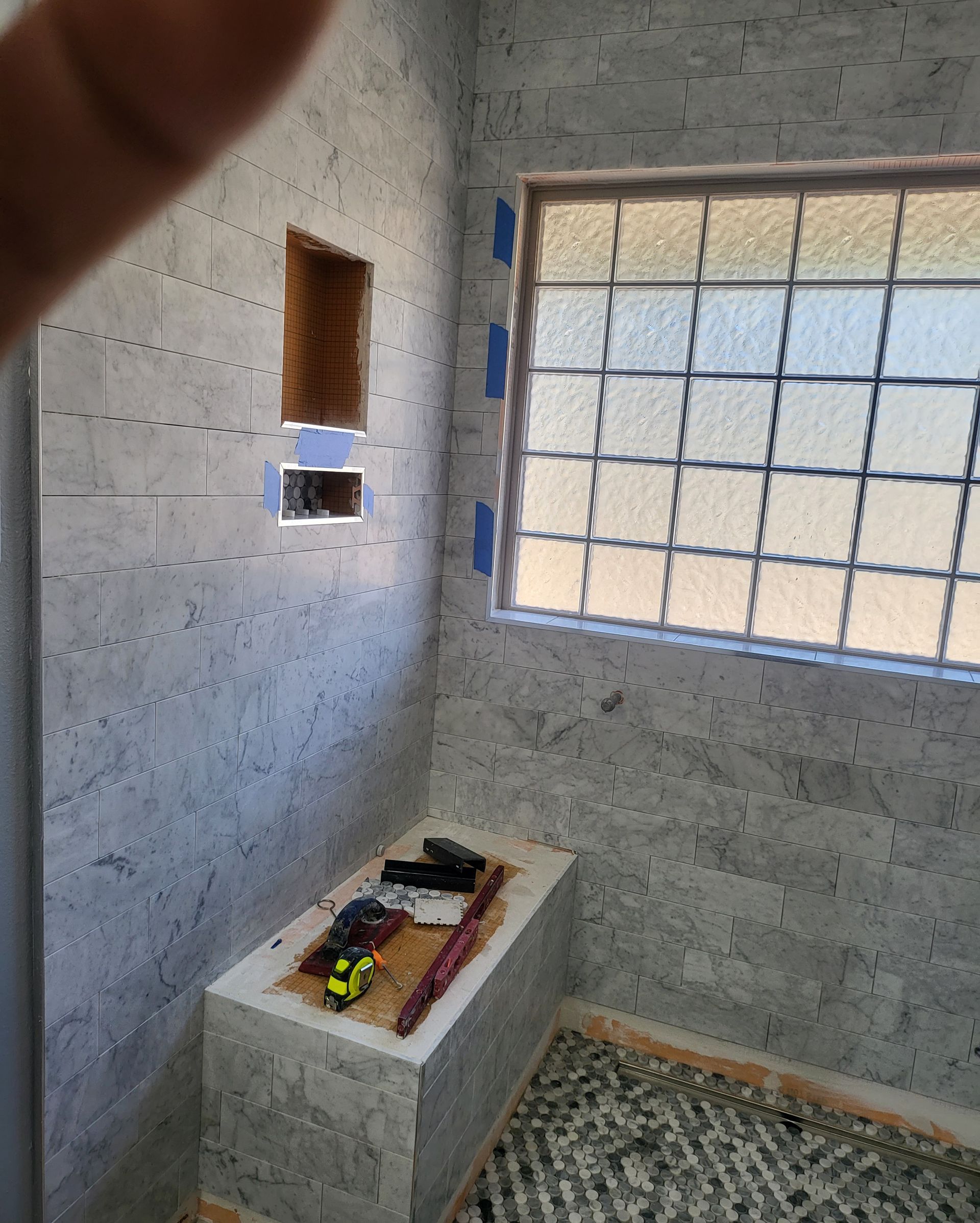 Small unfinished tiled bathroom with a window and a built-in bench holding tools and materials.