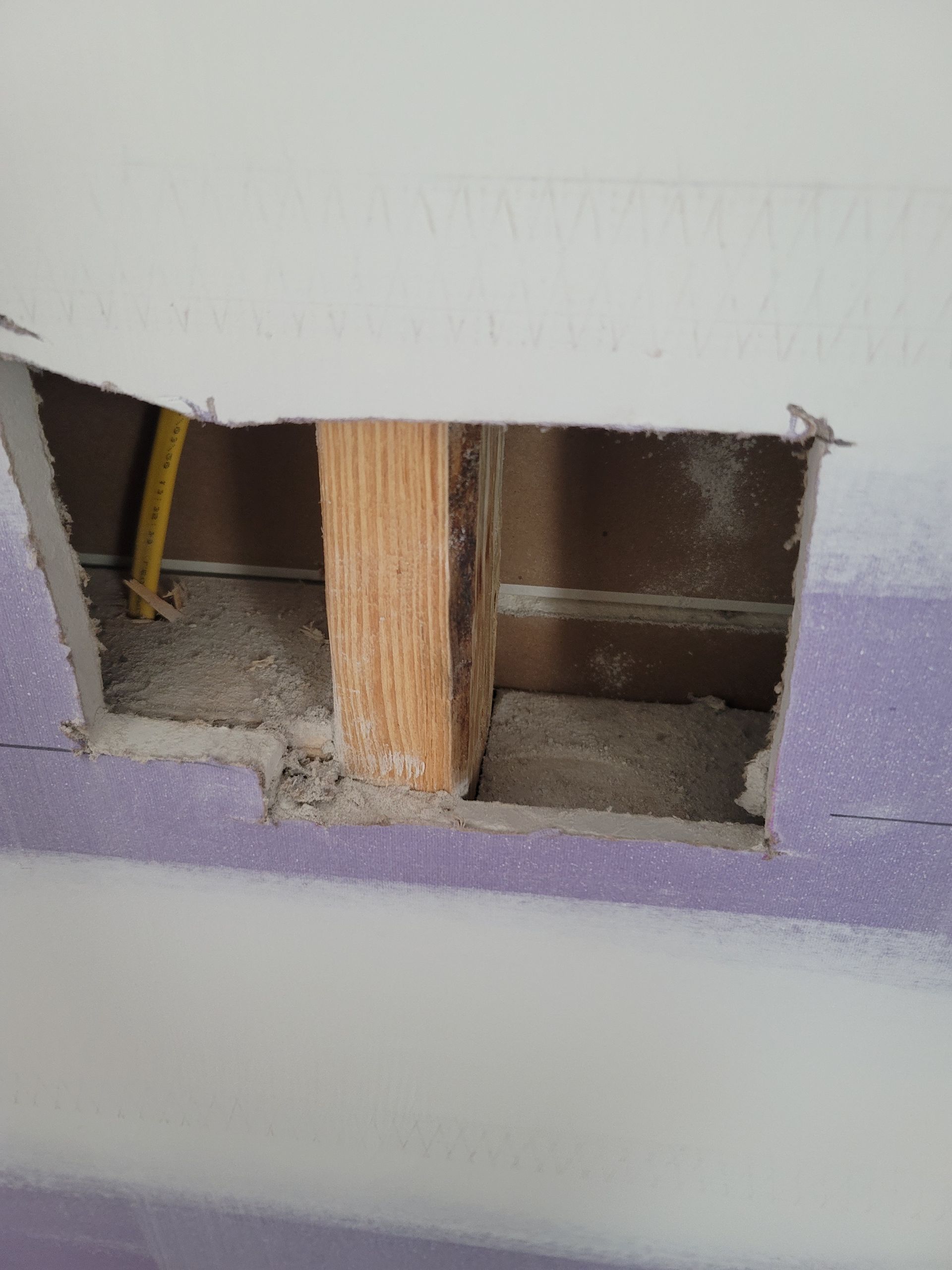 Damaged wall opening exposing wooden studs and wiring, with purple trim and white drywall edges