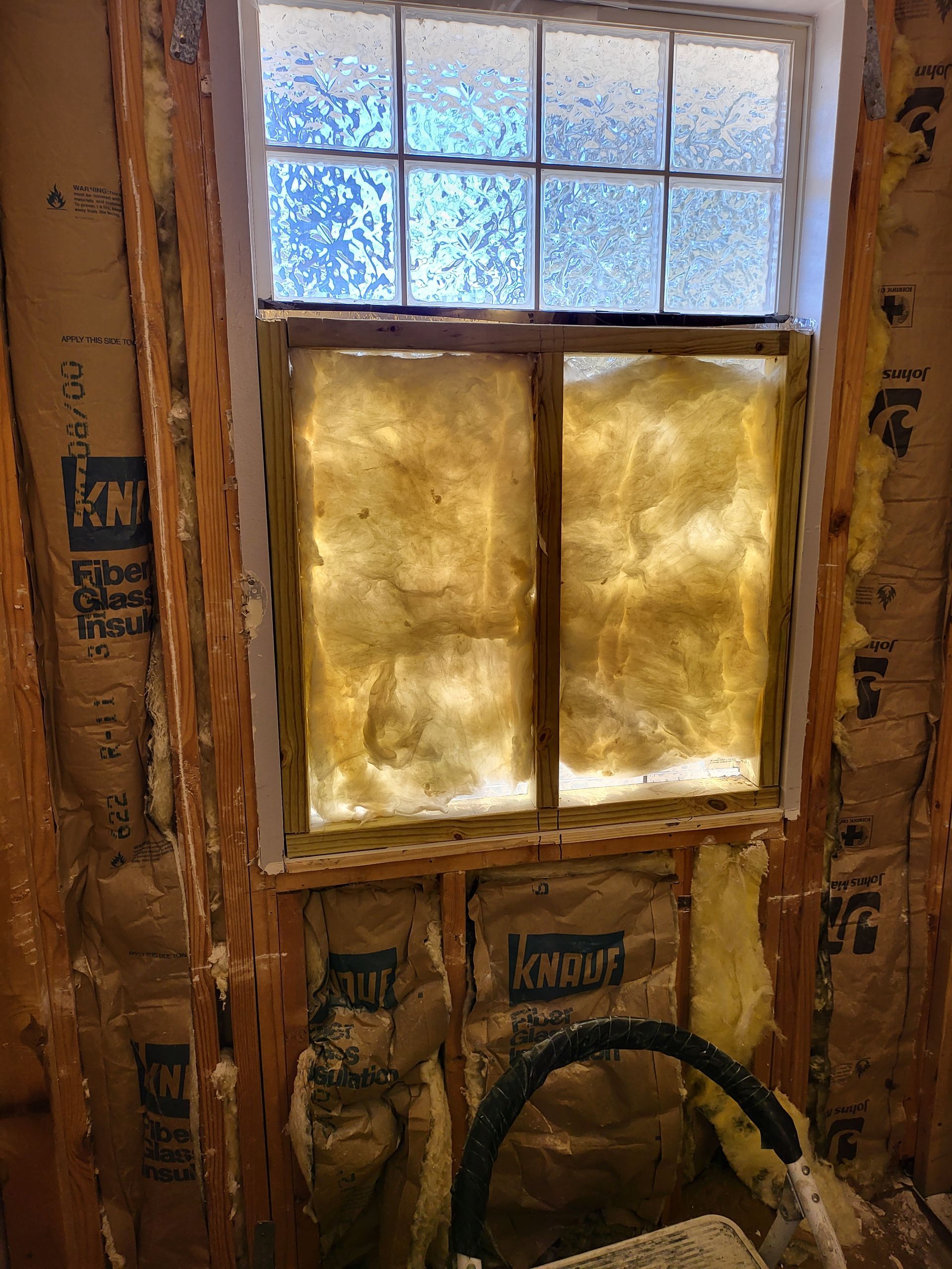 Insulation-covered wall with a small frosted window and exposed framing in a construction area