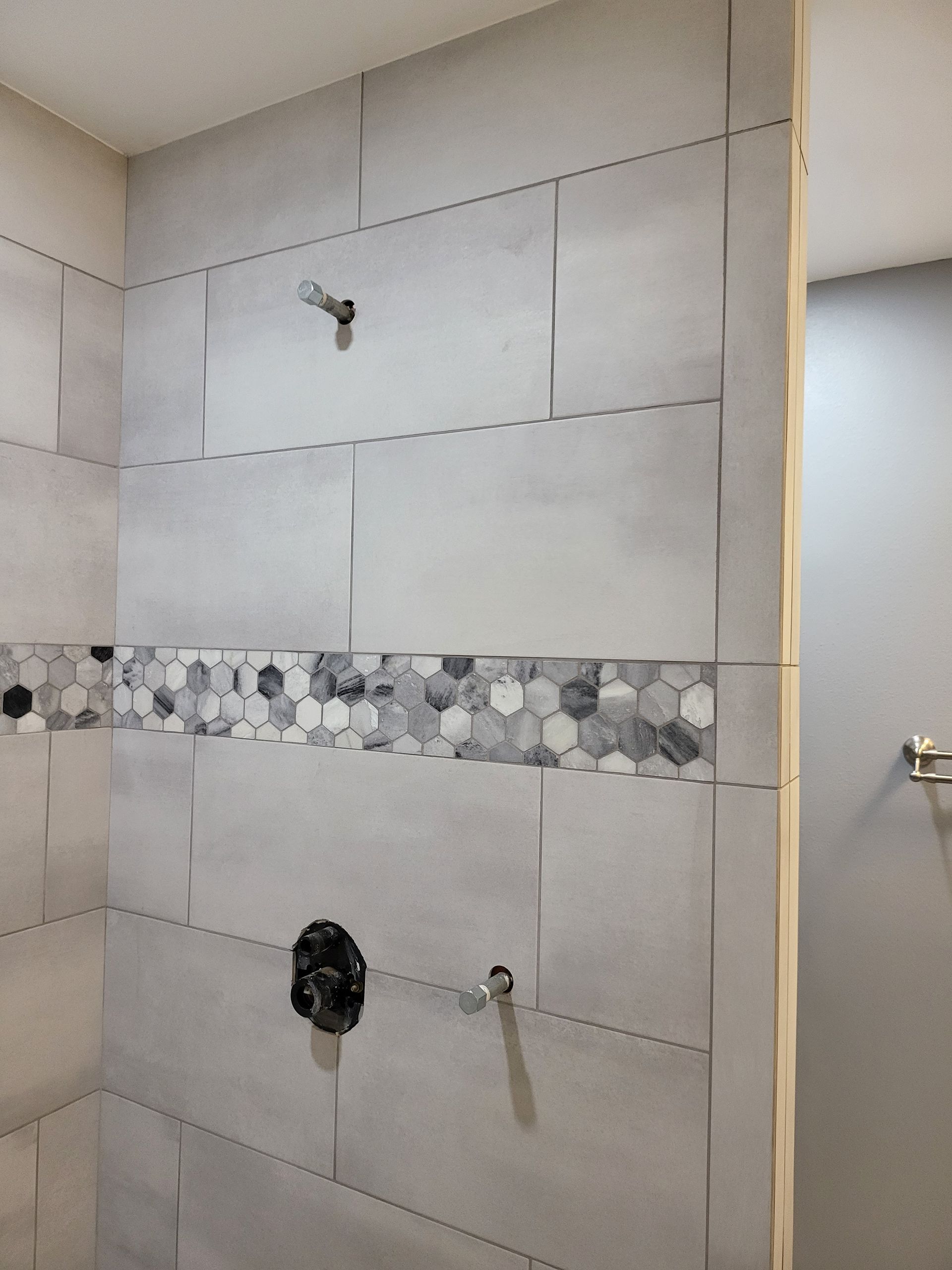 Bathroom shower wall with gray tiles and a pebble accent strip, showing plumbing fixtures installed.