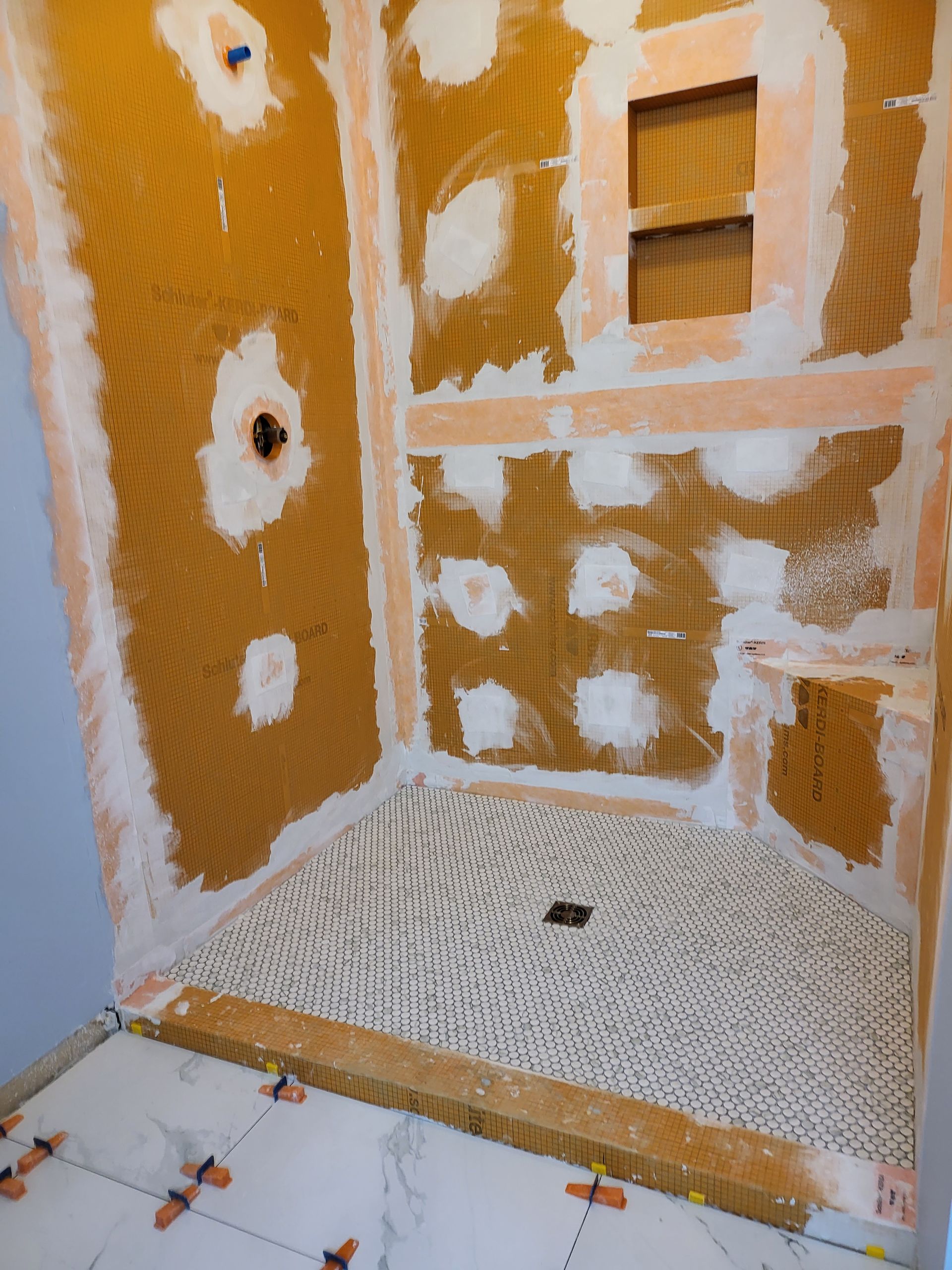 Partially tiled shower under renovation with orange waterproofing, white tile adhesive, and exposed wall niches
