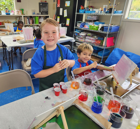 Two smiling students painting at a table in a classroom with colorful paints and easels.