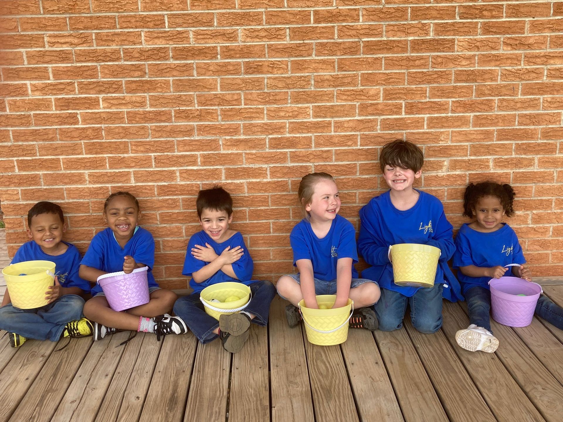 Children in blue shirts sit with colorful buckets against a brick wall, smiling.