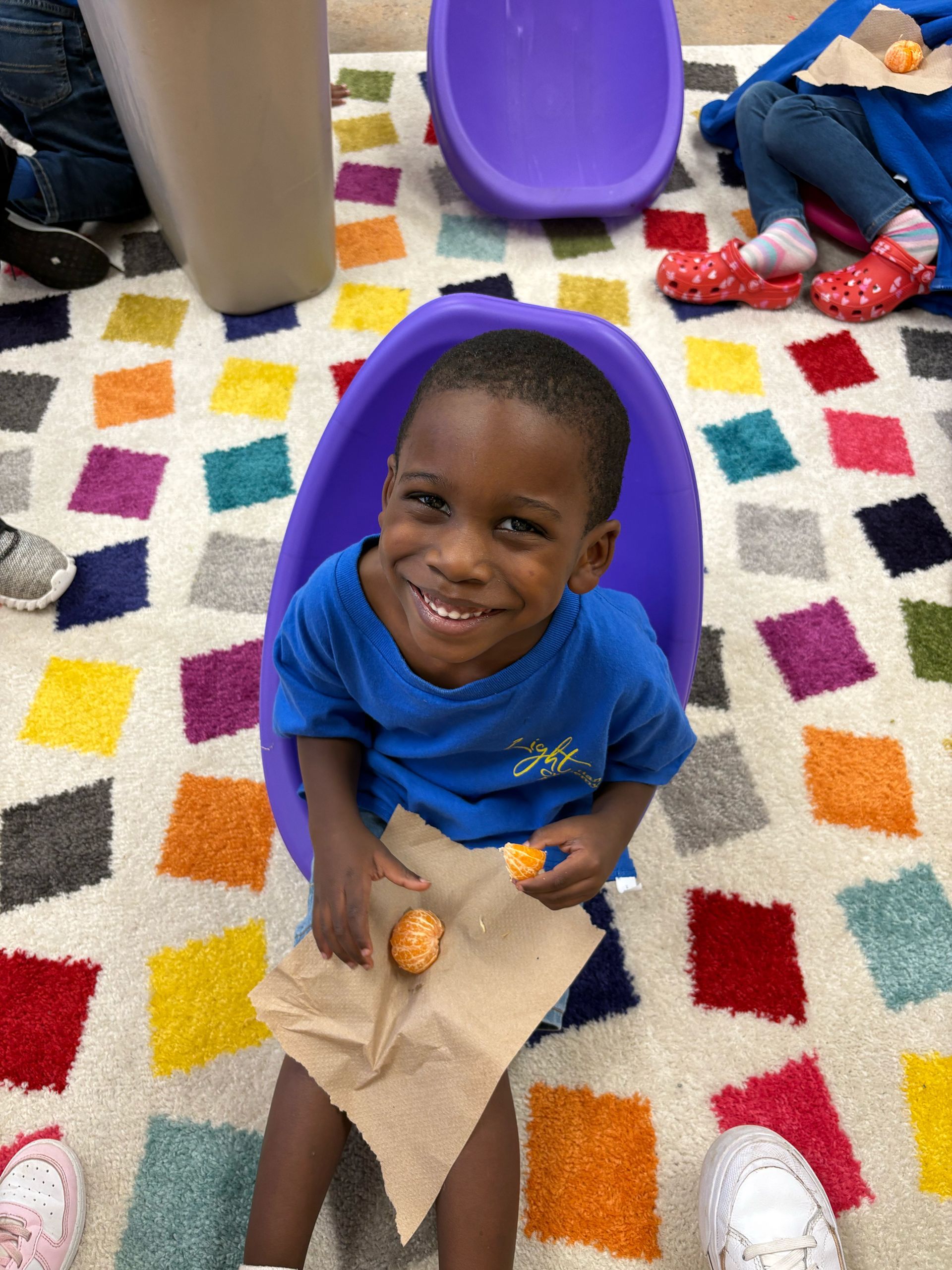 Child smiling, holding food, seated in purple chair on colorful rug.