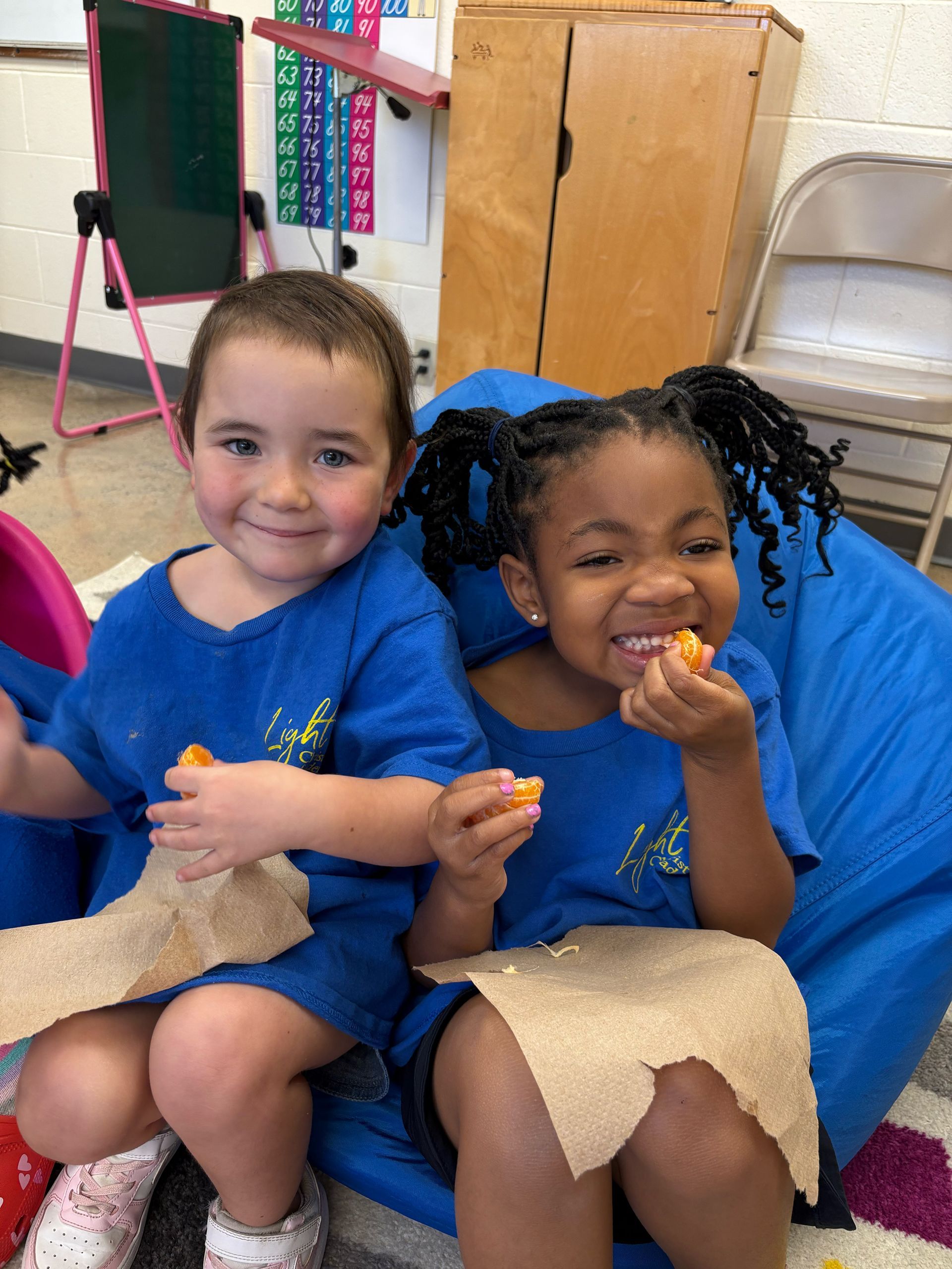 Two children in blue shirts smiling and eating snacks on a blue beanbag chair.