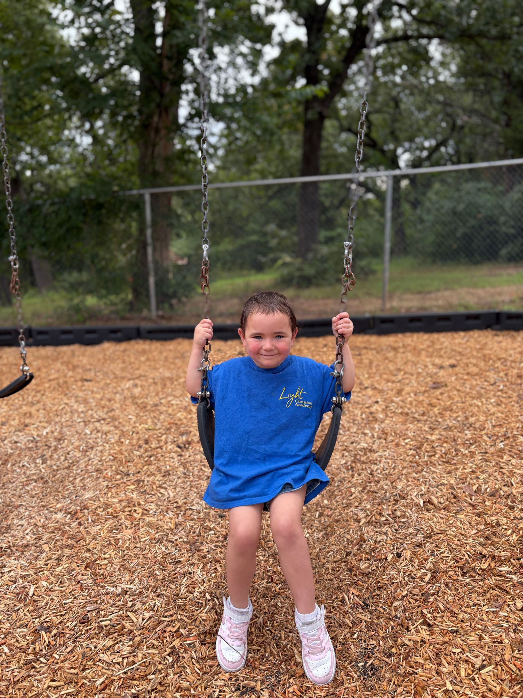 Child swinging at a playground, wearing blue shirt and white sneakers. Wood chips cover the ground.