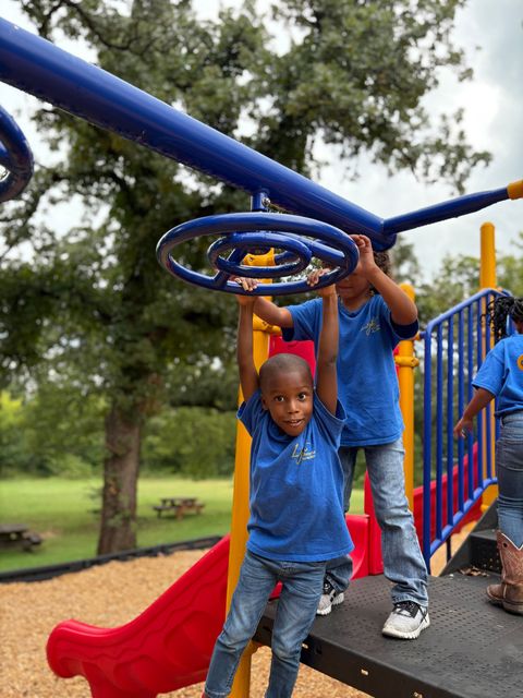 Children playing on a blue and red playground. One child hangs from a ring, another assists. Outdoors with trees.