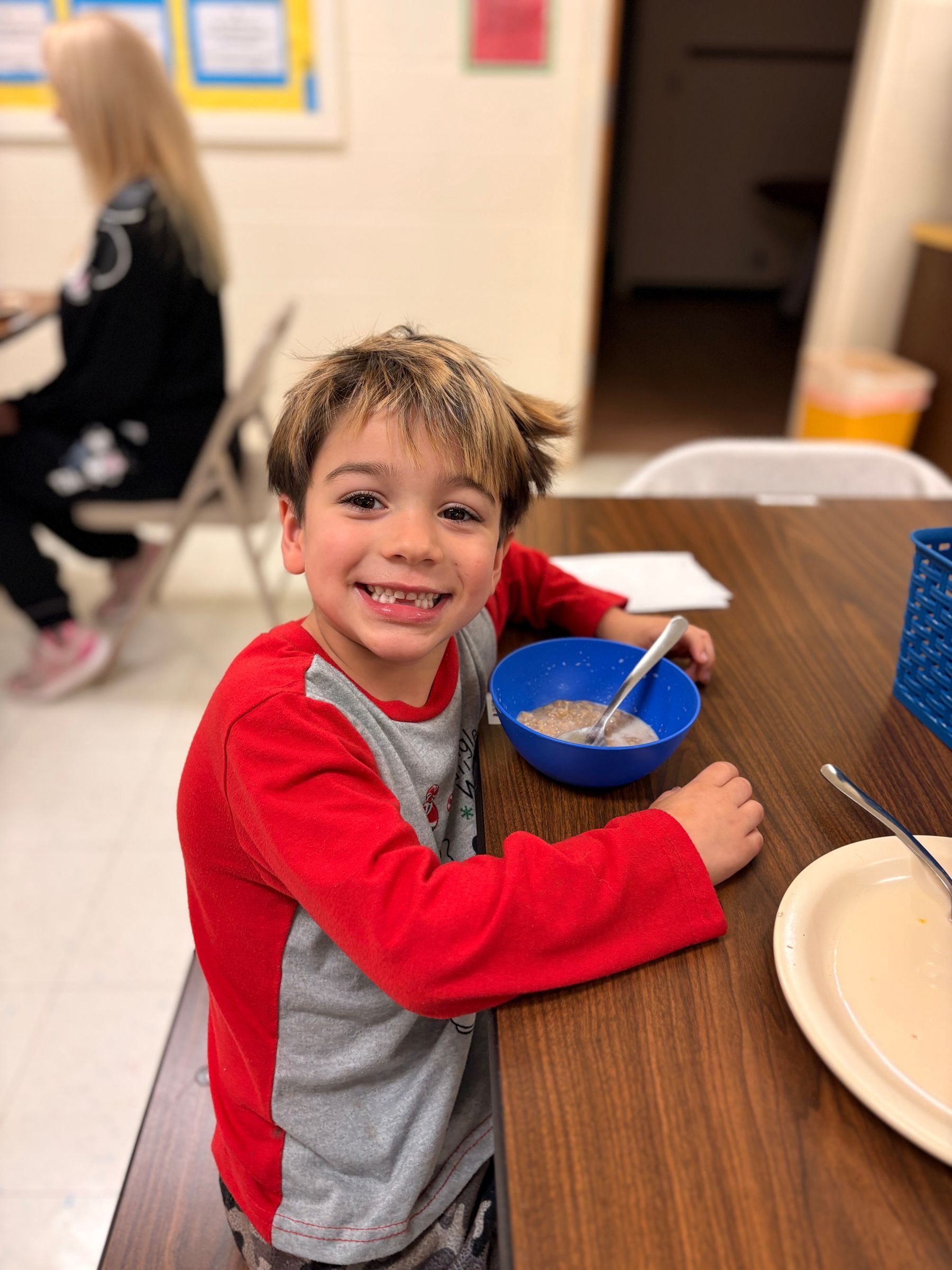 Boy smiling, eating cereal at a table. Red shirt, blue bowl. Woman in background.