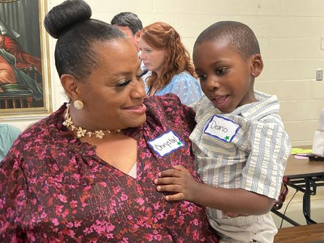 Woman with dark hair bun hugs a young boy. Both smile indoors, name tags visible.